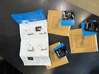 Na ciemnym stole leżą rozłożone materiały: biała instrukcja z niebieskimi elementami graficznymi i schematami, kilka żółtych kopert z niebieskimi naklejkami oraz małe czarne opakowania z nieczytelnym tekstem.