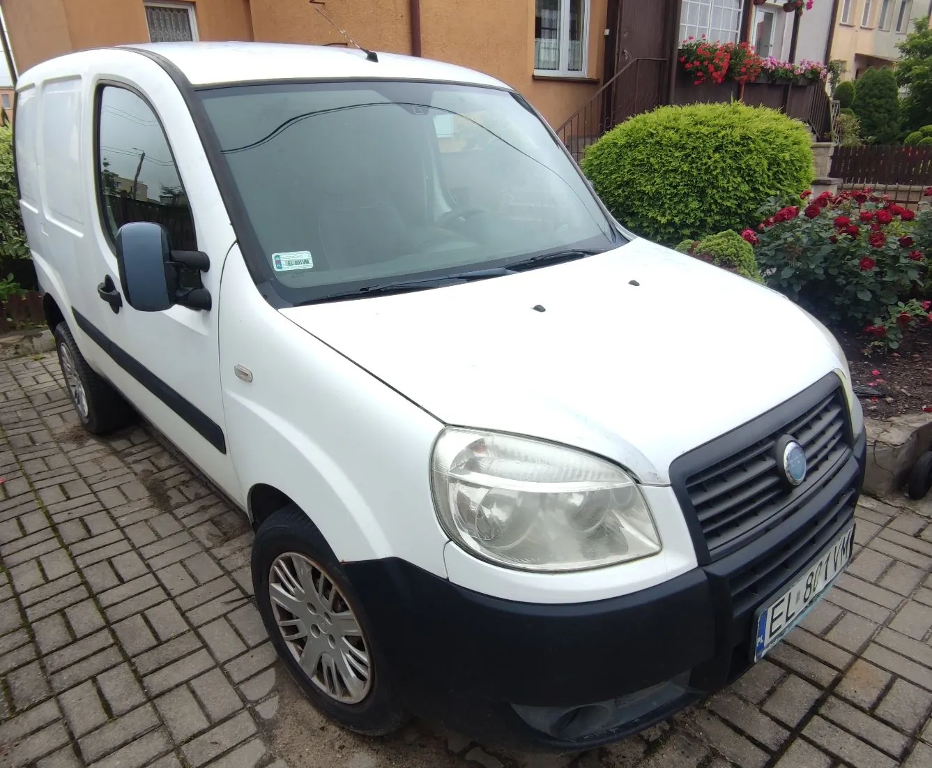 Biały samochód dostawczy marki Fiat Doblo zaparkowany na kostce brukowej przed domem. Pojazd jest widoczny z przodu i z boku. W tle znajduje się beżowy budynek z oknami, zielony krzew oraz krzewy z czerwonymi kwiatami.