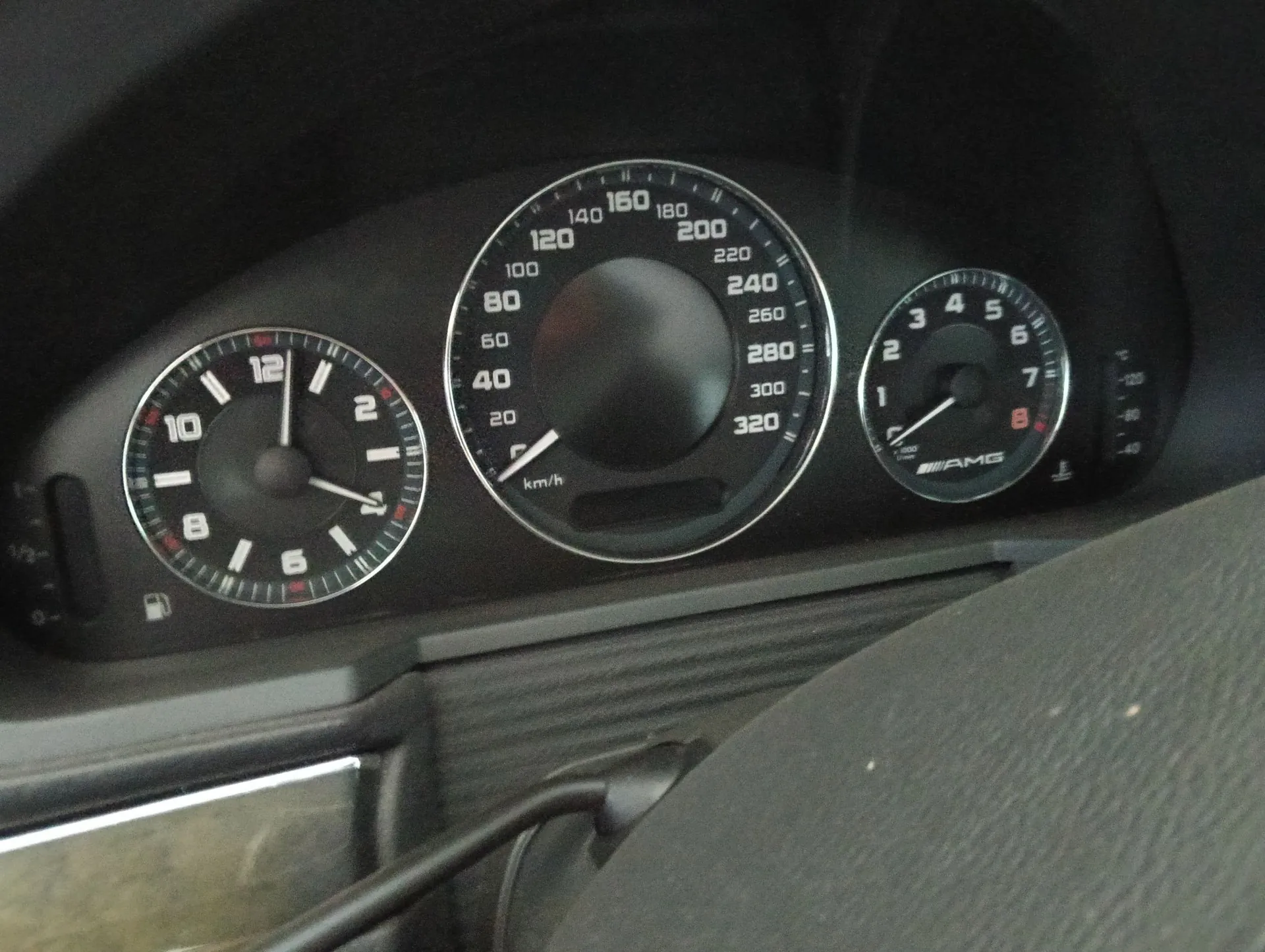 Zbliżenie na deskę rozdzielczą samochodu z trzema okrągłymi wskaźnikami. Centralny, największy licznik prędkości ma skalę od 0 do 320 km/h. Po lewej stronie znajduje się wskaźnik paliwa i temperatury, a po prawej obrotomierz z logo AMG na dole. Wskaźniki mają białe cyfry i czerwone akcenty na czarnym tle.