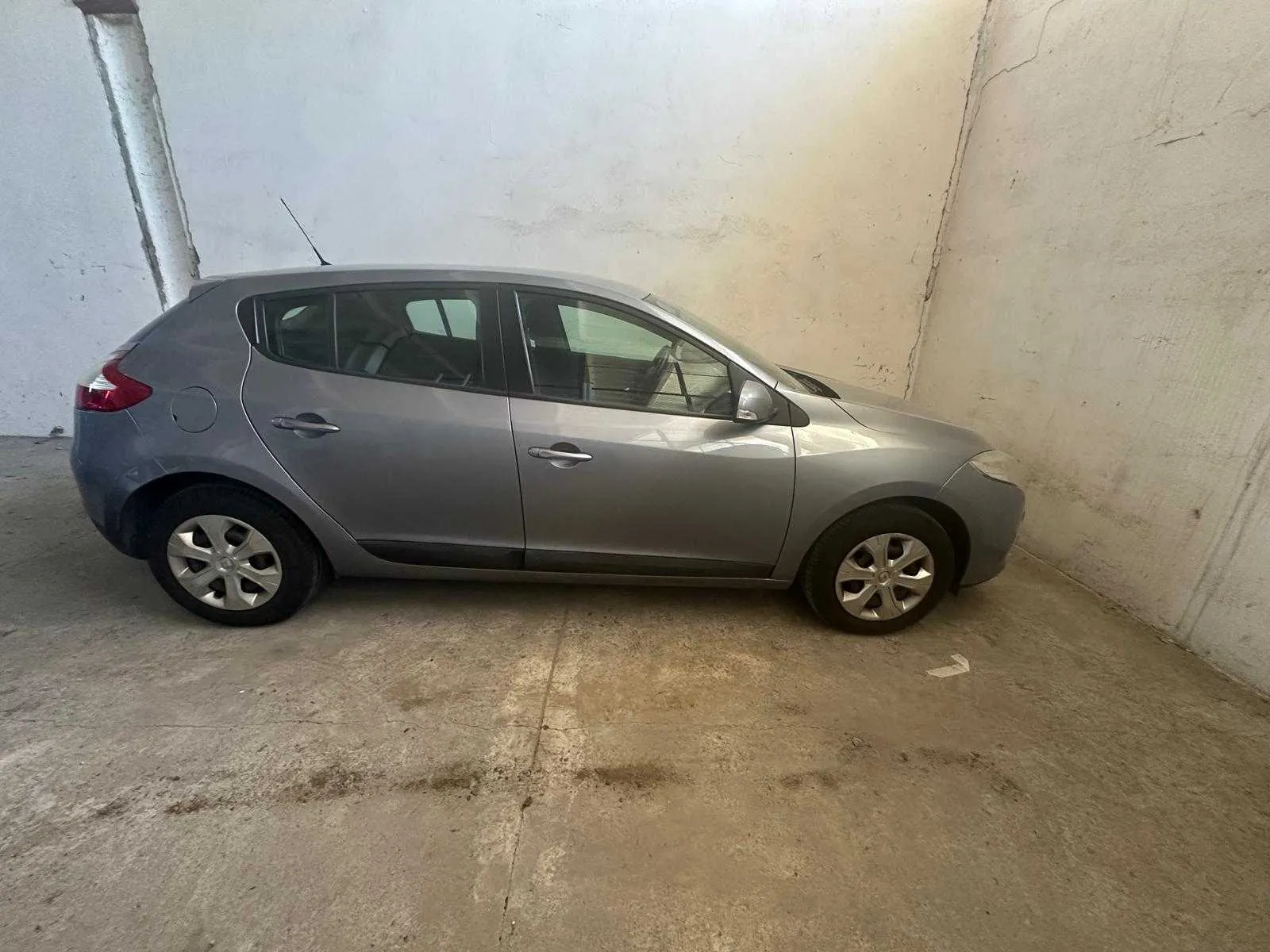 Srebrny samochód osobowy typu hatchback zaparkowany w pomieszczeniu z betonowymi ścianami i podłogą. Widok z boku, cztery koła, czerwone światło tylne, antena na dachu. Ściany mają pęknięcia i ślady zużycia.