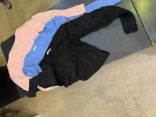 Trzy swetry w kolorach różowym, niebieskim i czarnym ułożone na ciemnej powierzchni. W tle widoczna betonowa podłoga oraz stopa osoby w białym obuwiu w lewym dolnym rogu.