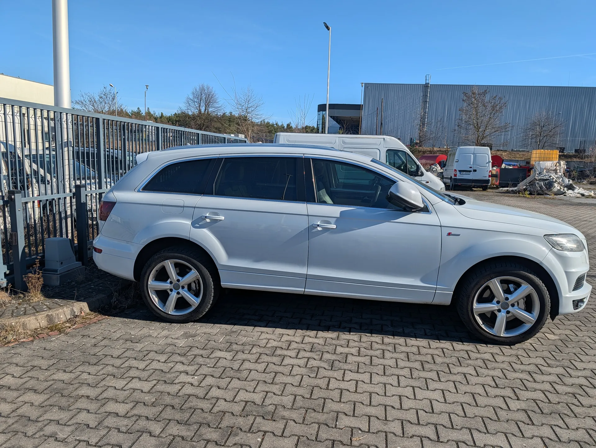 Srebrny samochód typu SUV zaparkowany na brukowanej powierzchni. W tle metalowe ogrodzenie, białe furgony, przemysłowa hala z metalową elewacją, suche drzewa oraz jasne niebo bez chmur. Samochód znajduje się w profilu, widoczne są cztery koła, lusterka boczne i okna. Na tle również słupy oświetleniowe i stosy materiałów.