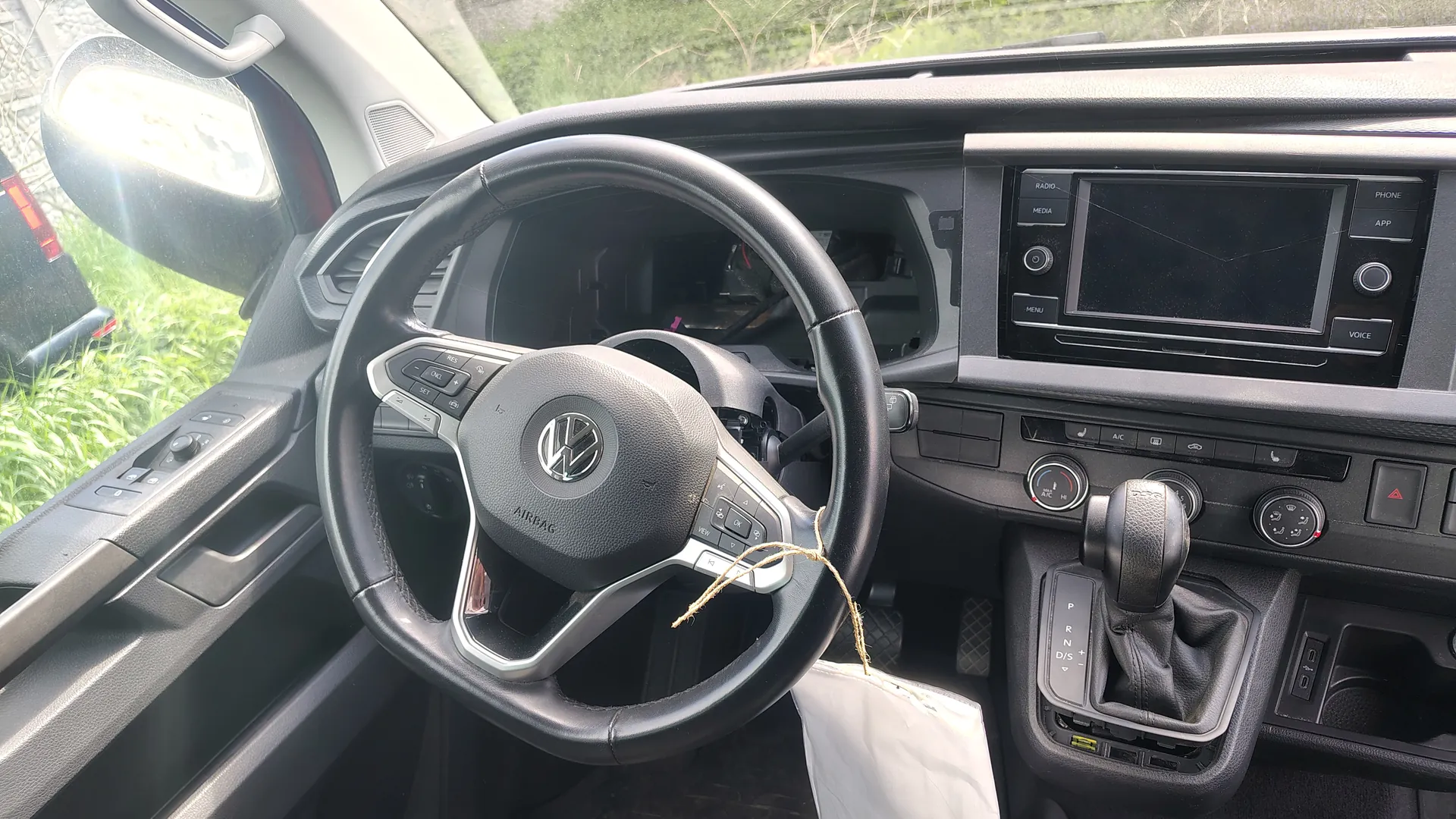 Wnętrze samochodu marki Volkswagen z widoczną kierownicą z logo VW i napisem AIRBAG. Na kierownicy znajdują się przyciski sterujące. W centrum deski rozdzielczej ekran multimedialny z przyciskami RADIO, MEDIA, PHONE, APP, VOICE oraz przyciskiem MENU. Poniżej ekranu widoczne są pokrętła klimatyzacji i dźwignia biegów z oznaczeniami P, R, N, D/S. Po lewej stronie kierownicy lusterko boczne i fragment drzwi z przyciskami. Na kierownicy zawieszona jest biała torba przywiązana sznurkiem. Za przednią szybą widoczna zielona trawa i fragment innego pojazdu.