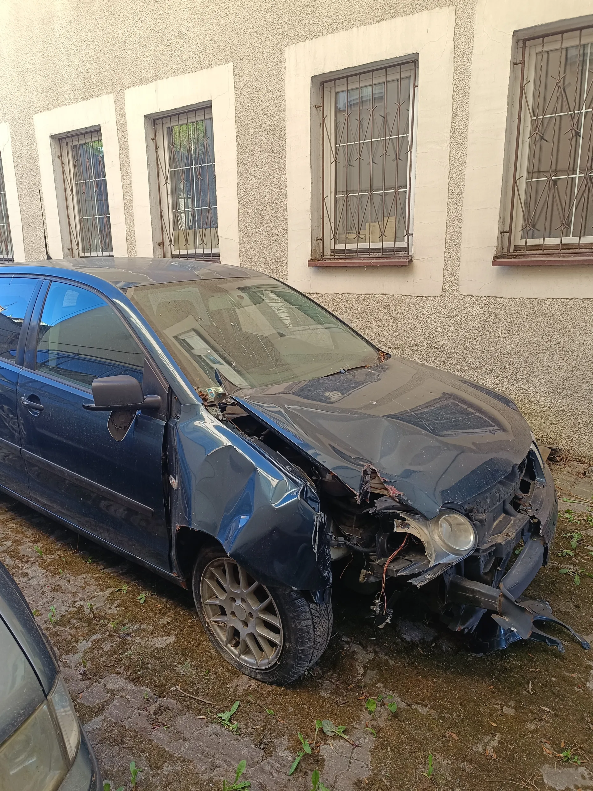 Zniszczony niebieski samochód osobowy zaparkowany przed budynkiem o szorstkiej, beżowej elewacji. Widoczne są cztery okna z metalowymi kratami i białymi obramowaniami. Przednia część pojazdu jest silnie uszkodzona: zdeformowany kapot, zniszczony zderzak, odsłonięte przewody i uszkodzone reflektory. Samochód stoi na brudnej, częściowo porośniętej trawą nawierzchni. W lewym dolnym rogu widoczna jest część innego pojazdu.