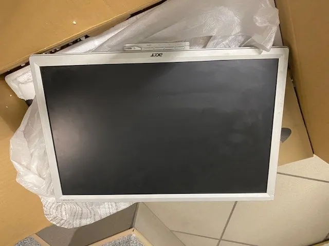 Monitor Acer o czarnym ekranie w srebrnej ramie, z widocznym logo 'Acer' po lewej stronie, umieszczony w kartonowym pudełku z białą folią ochronną. W tle widoczne są płytki podłogowe i części kartonów.
