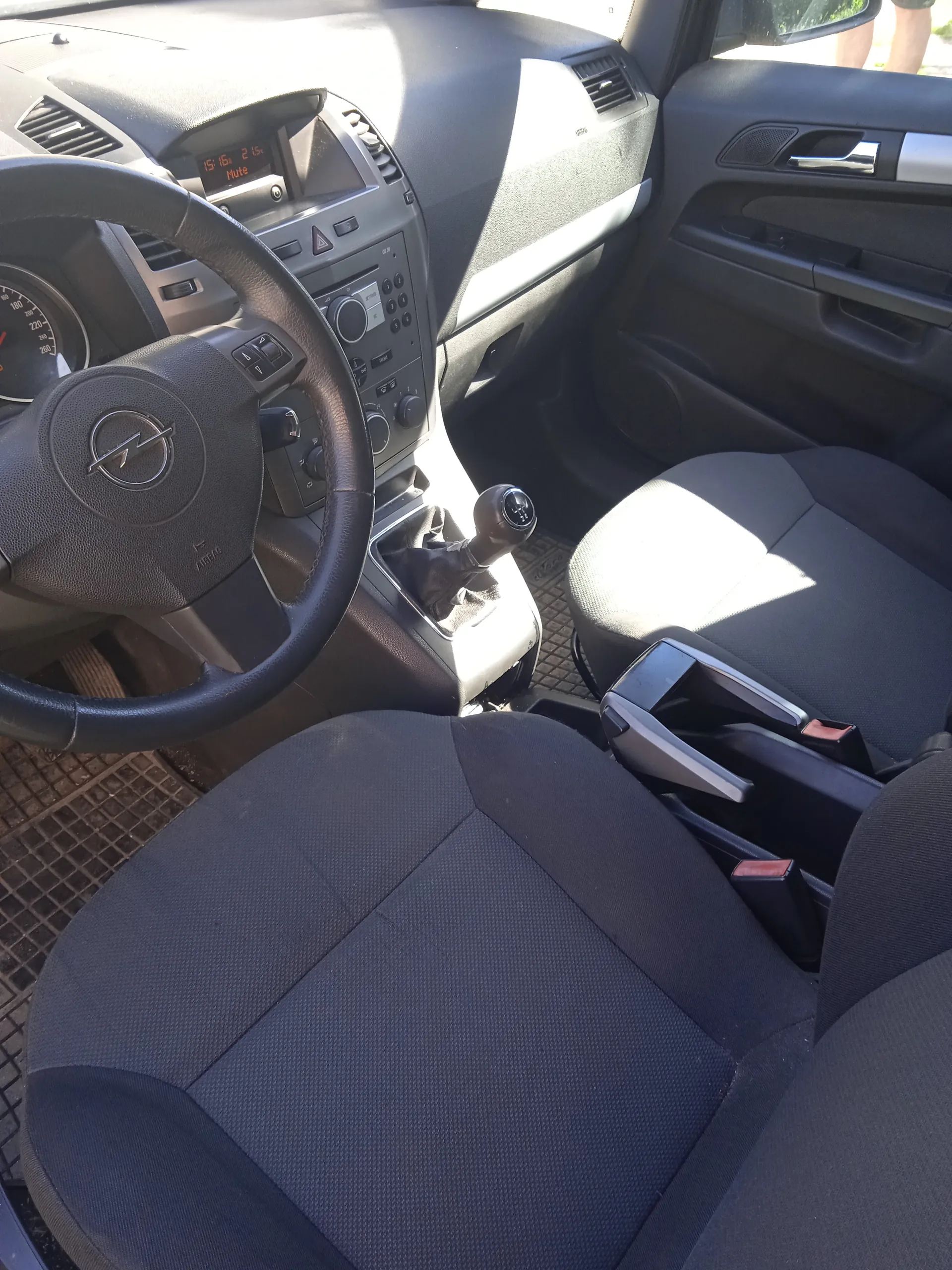 Wnętrze samochodu z widocznym kierownicą z logo Opel, czarnymi siedzeniami, deska rozdzielcza z wyświetlaczem cyfrowym pokazującym godzinę 15:16 i napisem Mute, przyciskami sterującymi, dźwignią zmiany biegów oraz drzwiami pasażerskimi z uchwytem i sterowaniem szybą. W tle widoczne są wentylacyjne otwory i fragment podłogi z matą.