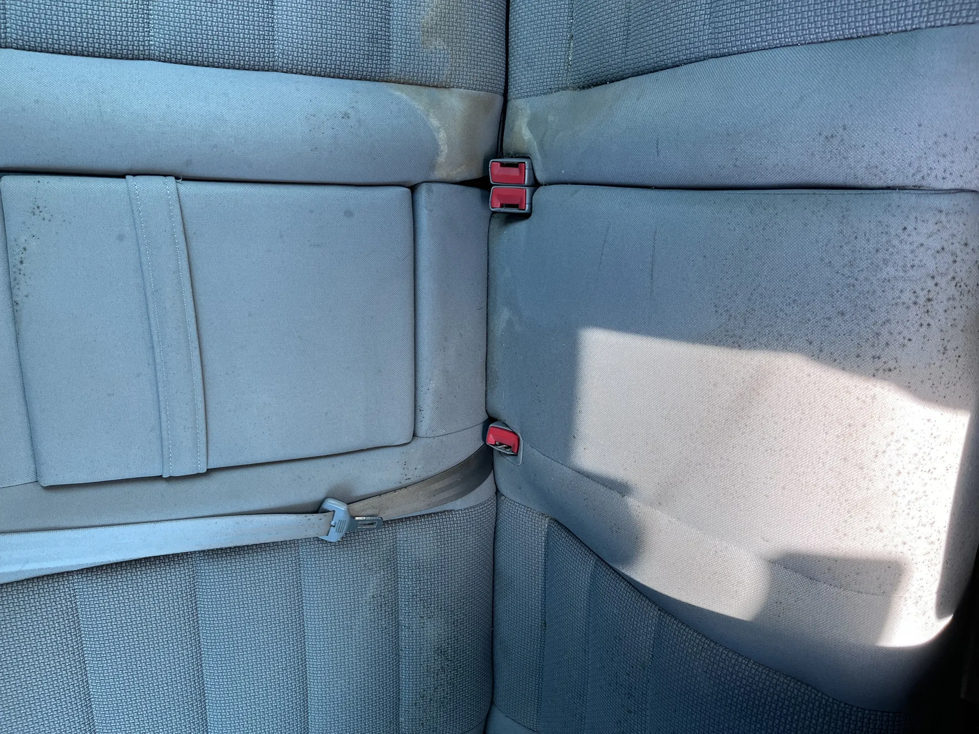 Wnętrze samochodu z widokiem na tylne siedzenia tapicerowane szarym materiałem. Widoczne są dwa czerwone zatrzaski pasów bezpieczeństwa oraz pasy bezpieczeństwa, z których jeden jest przypięty. Na siedzeniach znajdują się plamy i zabrudzenia. W prawej części obrazu widoczne jest oświetlenie słoneczne rzucające cień na siedzenie.