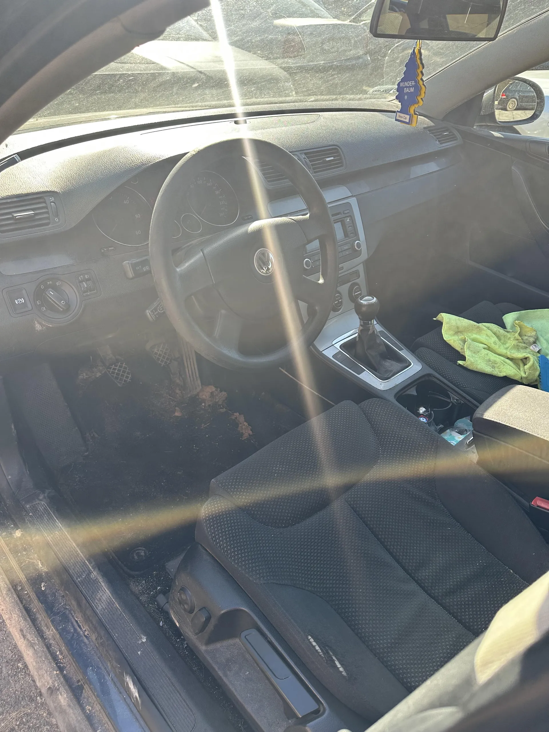 Wnętrze samochodu marki Volkswagen z widocznym kierownicą z logo VW, deska rozdzielcza z licznikami, dźwignią zmiany biegów i zielonym ręcznikiem na siedzeniu pasażera. Na podłodze widoczne są suche liście. W lusterku wstecznym wiszący niebieski odświeżacz powietrza z napisem "WUNDER-BAUM". Drzwi kierowcy są otwarte, a przez przednią szybę widać inny samochód na zewnątrz. Słoneczne promienie wpadają do wnętrza.