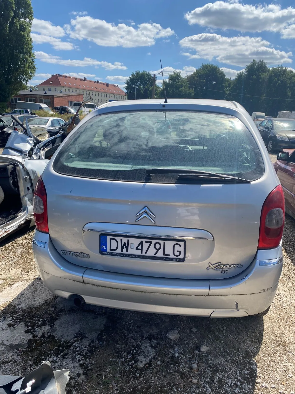 Srebrny samochód marki Citroën Xsara widziany od tyłu. Pojazd posiada tablicę rejestracyjną o numerze DW 479LG. Samochód znajduje się na piaszczystym terenie obok innych uszkodzonych wraków aut. W tle widoczne są zielone drzewa, budynek mieszkalny oraz niebieskie niebo z białymi chmurami.