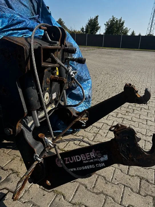Zbliżenie na ciężki, metalowy element maszyny rolniczej lub przemysłowej w kolorze czarnym, z widocznymi śladami rdzy i zużycia. Urządzenie jest zabezpieczone stalowymi linami i znajduje się na kostce brukowej. W tle widoczna jest niebieska plandeka oraz ciemny płot i drzewa pod jasnym niebem. Na dolnej części metalowej ramy widnieje napis: 'ZUIDBERG FROM FINE LINE SYSTEMS www.zuidberg.com'.