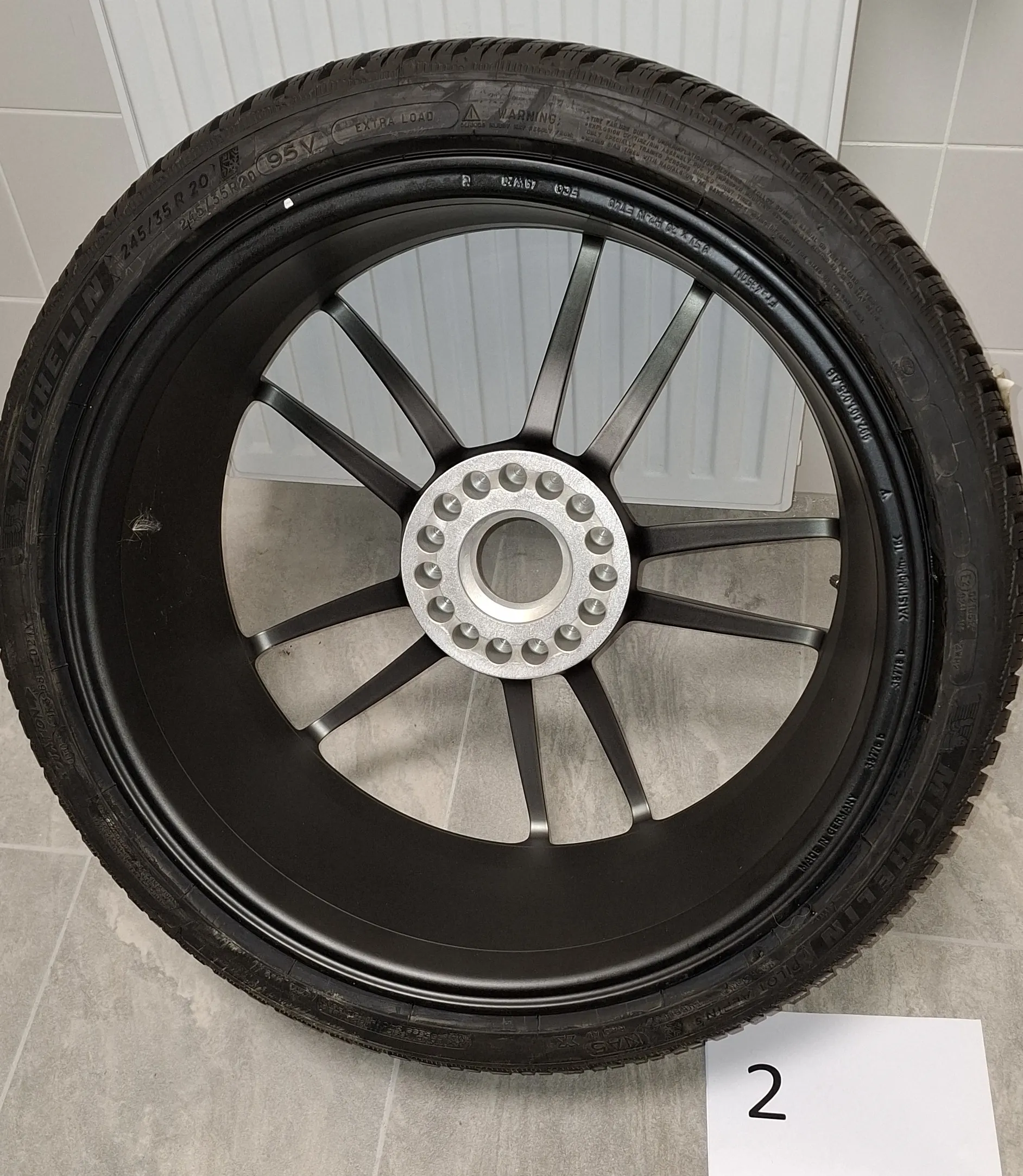 Czarna felga samochodowa z założoną oponą marki Michelin, oparta o biały grzejnik na jasnym tle. Felga ma wiele cienkich ramion i srebrny środek z otworami na śruby. Na boku opony widoczny jest napis 'MICHELIN' oraz rozmiar '245/35 R 20 95V EXTRA LOAD'. W prawym dolnym rogu zdjęcia znajduje się biała kartka z czarną cyfrą '2'.