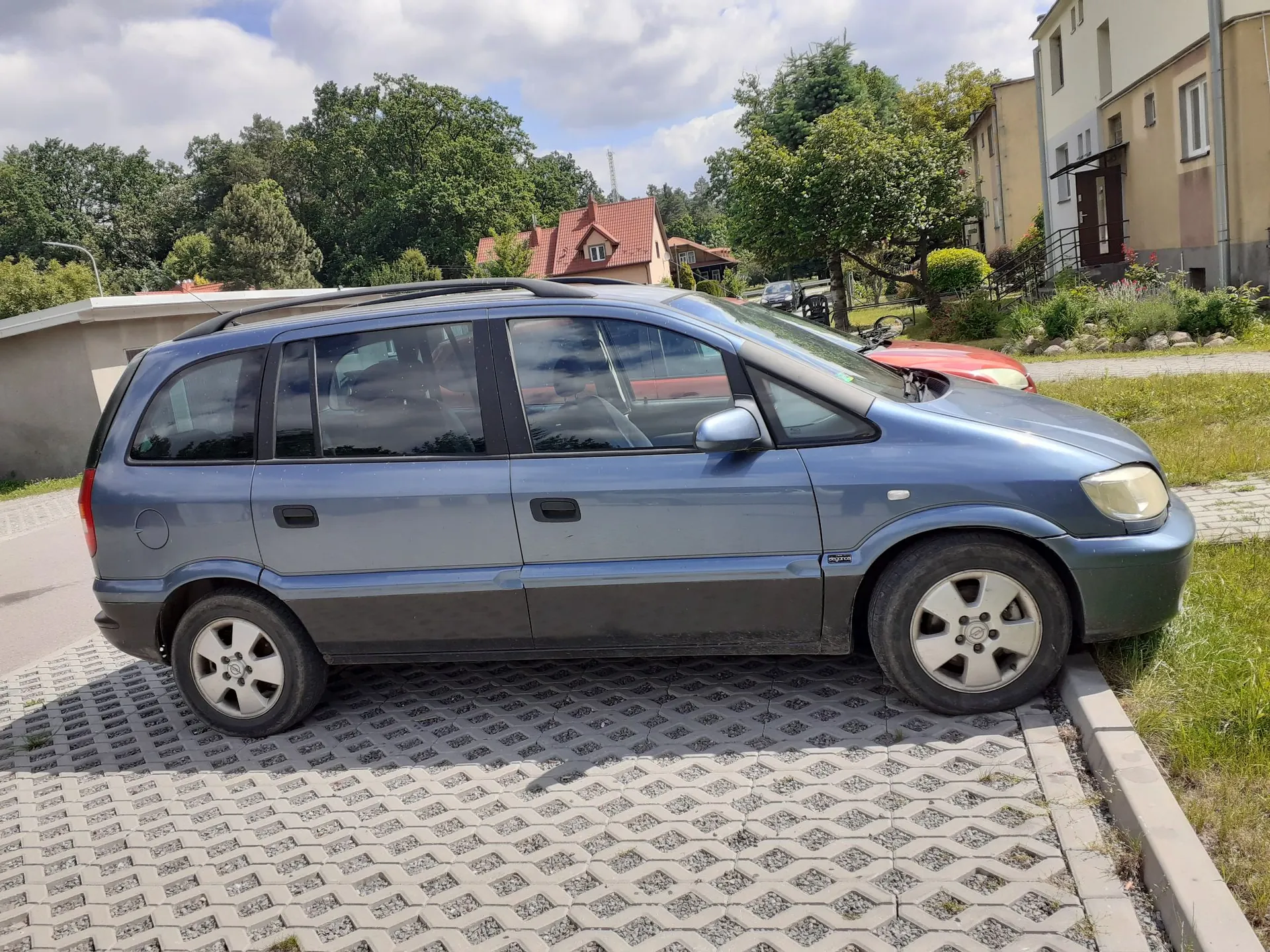 Niebieski samochód typu minivan zaparkowany bokiem na placu z betonowej kratki trawowej. W tle widoczne są zielone drzewa, dom z czerwonym dachem oraz fragmenty budynków mieszkalnych pod jasnym, lekko zachmurzonym niebem.