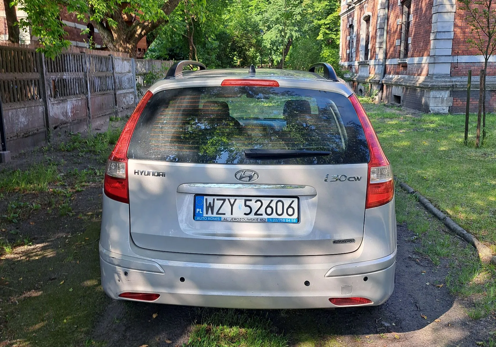 Srebrny samochód marki Hyundai i30 cw zaparkowany tyłem do widza na trawiastym podjeździe. Na klapie bagażnika widnieje napis HYUNDAI po lewej stronie, logo marki na środku oraz oznaczenie i30 cw po prawej stronie. Samochód posiada polską tablicę rejestracyjną o numerze WZY 52606. W tle widoczny jest drewniany płot, zielone drzewa oraz fragment ceglanego budynku.