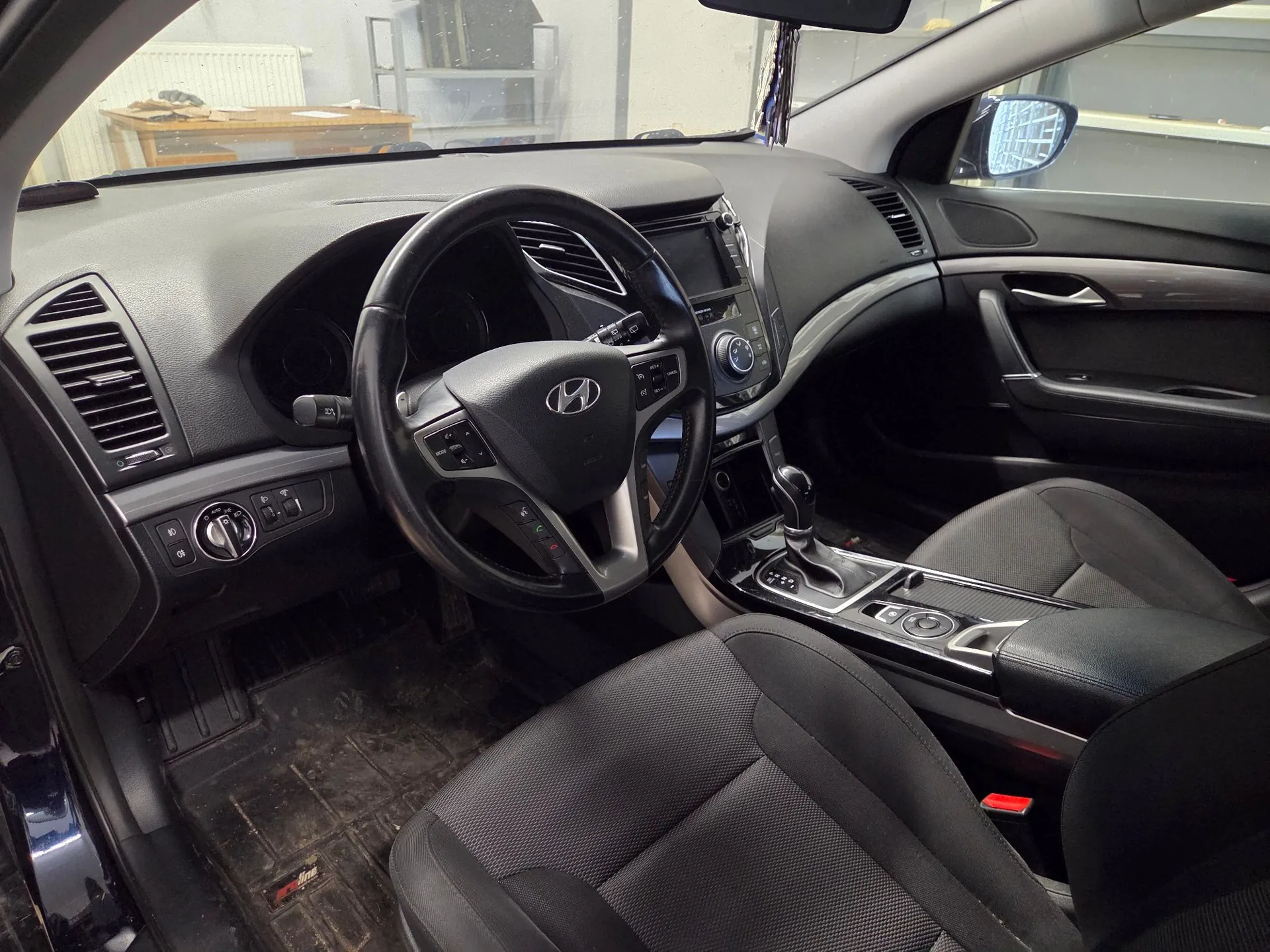 Wnętrze samochodu marki Hyundai widziane z perspektywy pasażera. Widoczna jest czarna kierownica z logo producenta, deska rozdzielcza z nawiewami, konsola środkowa z automatyczną skrzynią biegów oraz dwa przednie fotele obite czarnym materiałem.
