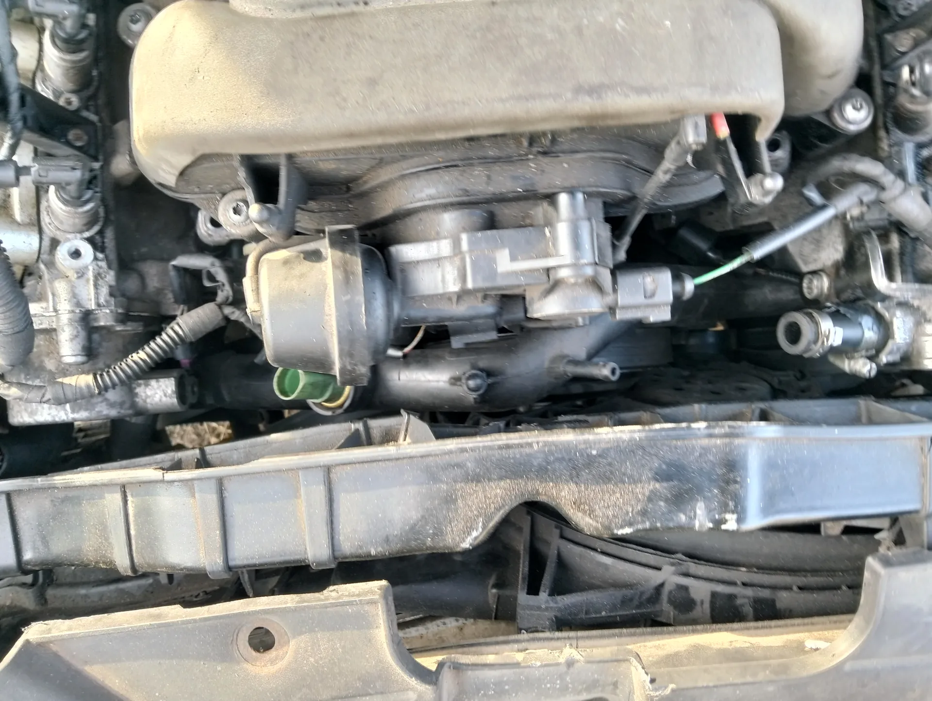 Zbliżenie na silnik samochodowy z widocznymi elementami takimi jak metalowe i plastikowe komponenty, przewody, węże oraz zielony korek. Silnik pokryty jest kurzem, a w tle znajdują się różne części mechaniczne i elektryczne. Brak czytelnego tekstu.