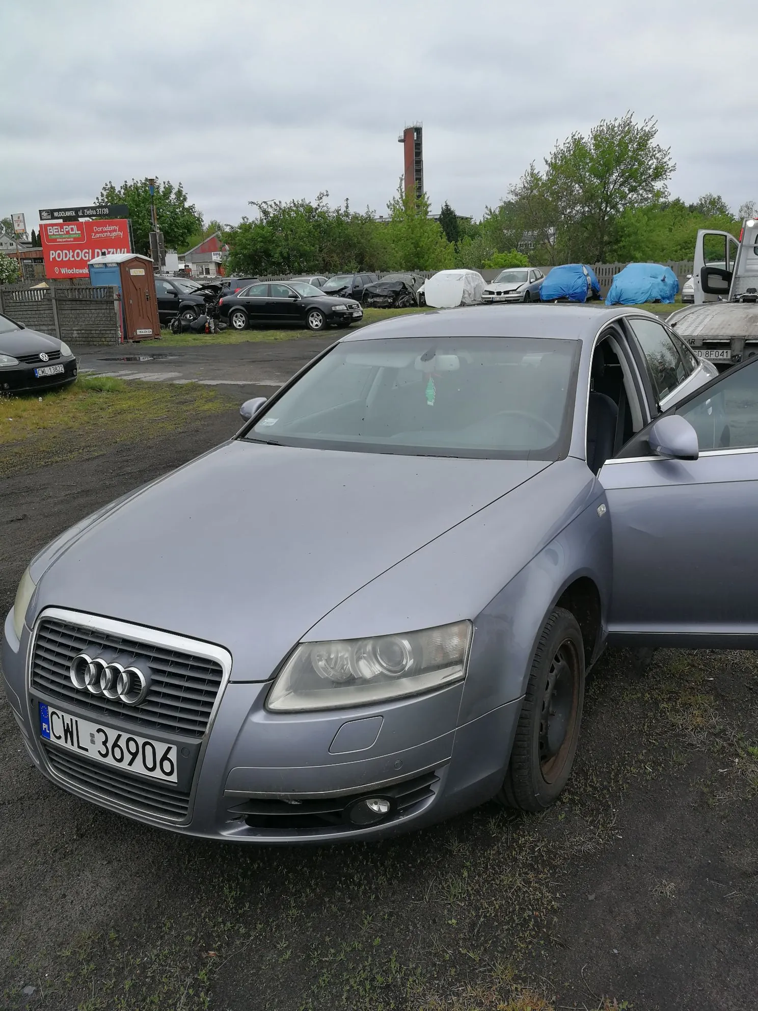 Srebrny samochód marki Audi zaparkowany na utwardzonym placu. Widoczna jest przednia część pojazdu z tablicą rejestracyjną o numerze CWL 36906. Drzwi pasażera są otwarte. W tle znajduje się parking z innymi samochodami, niebieskie plandeki oraz tablica reklamowa z napisami 'BELPOL' i 'PODŁOGI'. W oddali widoczny jest wysoki, wąski budynek przypominający wieżę.