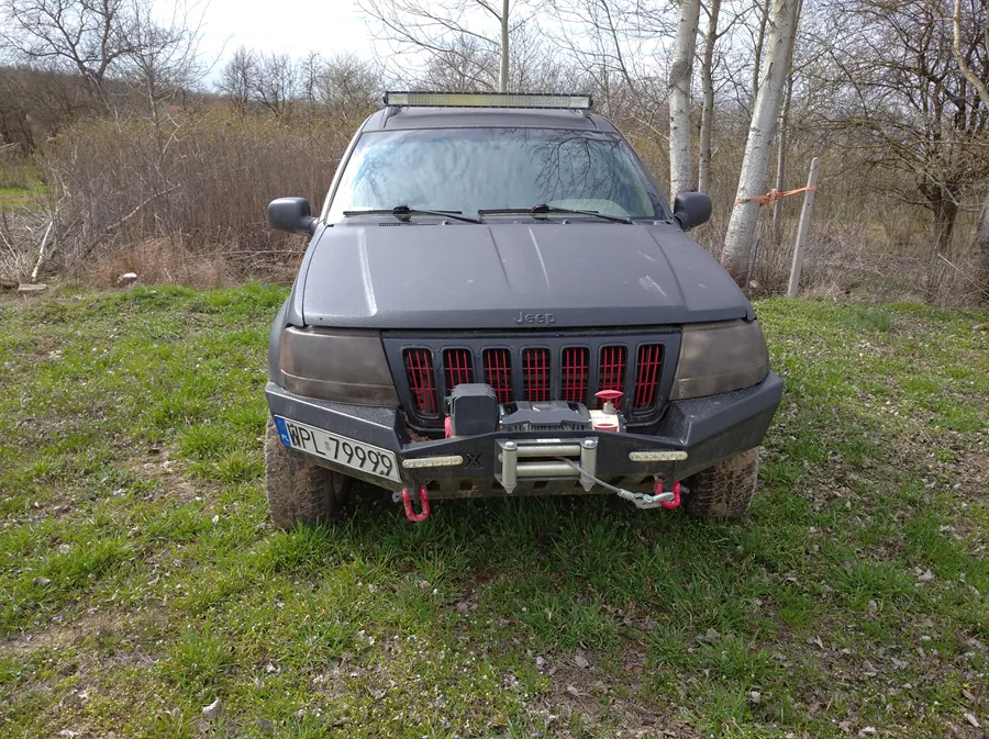 Ciemnoszary samochód terenowy Jeep z przodu, stojący na trawie. Widoczna tablica rejestracyjna HPL 79999, czerwone elementy na atrapie chłodnicy, wyciągarka z czerwonymi hakami na zderzaku oraz dachowy reflektor. Tło stanowią suche krzewy, drzewa bez liści i niebo. Samochód znajduje się na trawiastym terenie z widocznymi śladami opon.