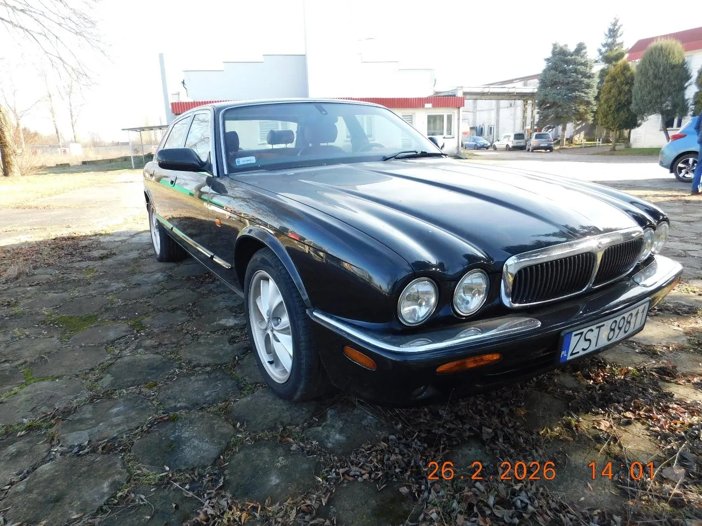 Czarny samochód marki Jaguar zaparkowany na brukowanej powierzchni z suchymi liśćmi. Widoczna tablica rejestracyjna ZST 89811 z oznaczeniem PL. W tle budynki z czerwonym dachem, inne pojazdy, drzewa oraz data i godzina 26.2.2026 14:01 umieszczone pomarańczowym kolorem w dolnej części obrazu.