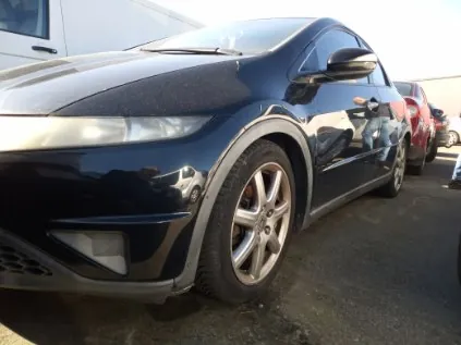 Czarny samochód osobowy typu hatchback, widok z przodu i boku, zaparkowany na asfaltowym parkingu. W tle widoczne są inne pojazdy, w tym czerwony samochód i biały van. Słoneczny dzień, odbicia na karoserii.