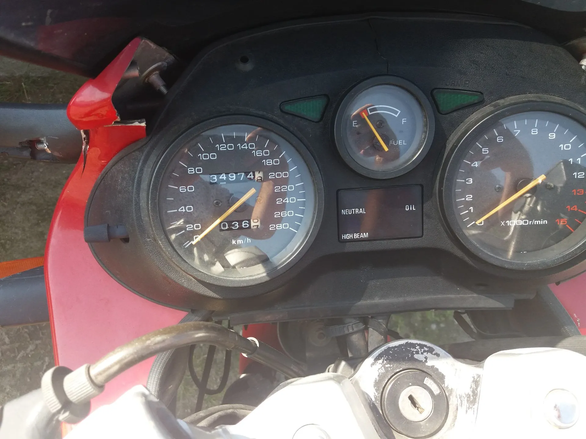 Zbliżenie na deskę rozdzielczą motocykla. Widoczny jest prędkościomierz z maksymalną wartością 280 km/h i przebiegiem 34974 km, wskaźnik poziomu paliwa (FUEL) oraz obrotomierz z oznaczeniem x1000r/min. Poniżej wskaźnika paliwa znajdują się kontrolki NEUTRAL, OIL oraz HIGH BEAM. Obudowa motocykla jest czerwona, a w dolnej części zdjęcia widoczna jest kierownica oraz stacyjka.