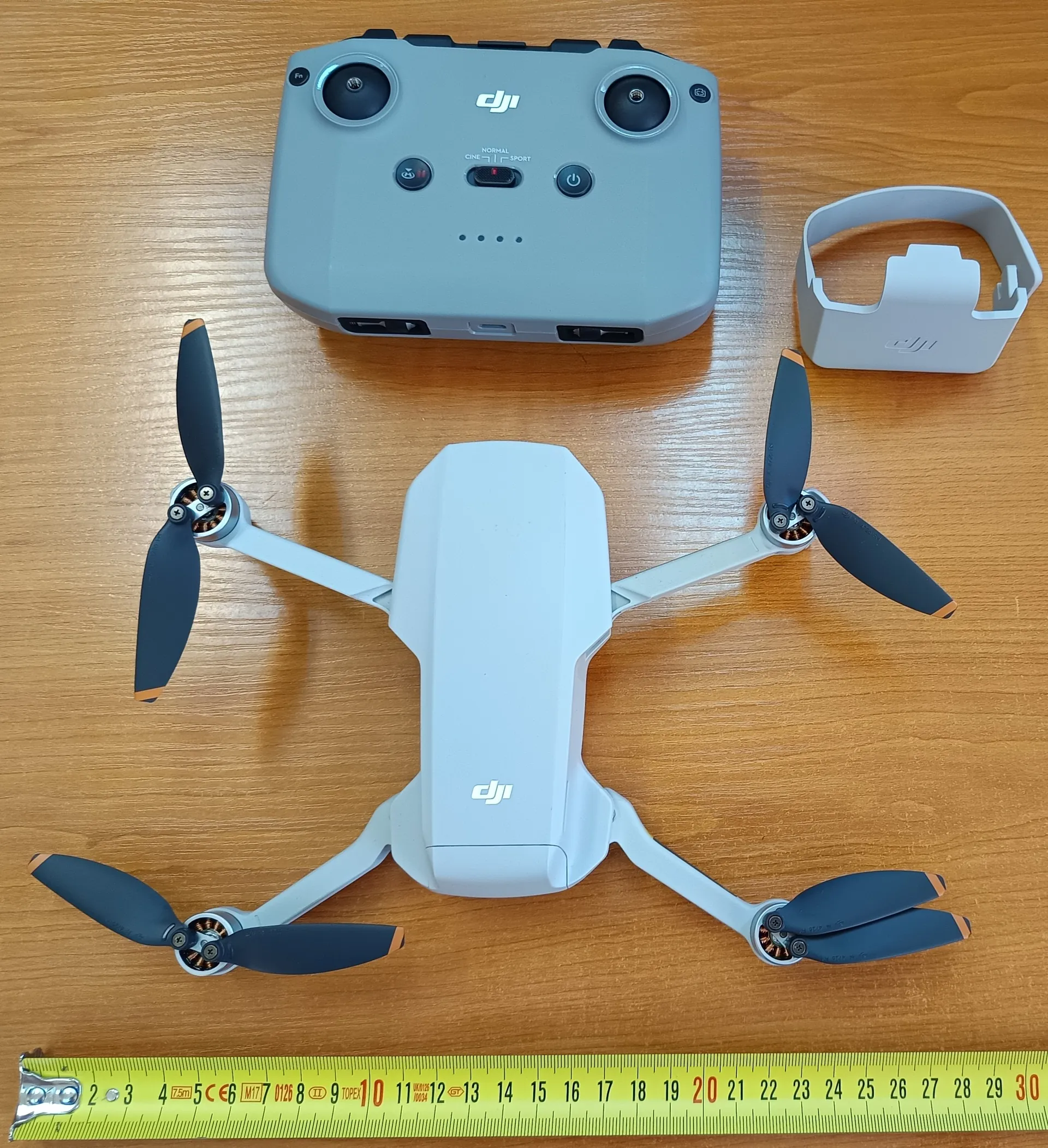 Na drewnianym stole umieszczono biały dron DJI z czterema czarnymi wirnikami o pomarańczowych końcówkach, szary pilot zdalnego sterowania z logo DJI i oznaczeniami trybów NORMAL, CINE, SPORT, szary uchwyt na telefon z logo DJI oraz żółtą taśmę mierniczą z czarnymi i czerwonymi oznaczeniami od 2 do 30. Wszystkie elementy są ułożone równolegle do krawędzi obrazu.