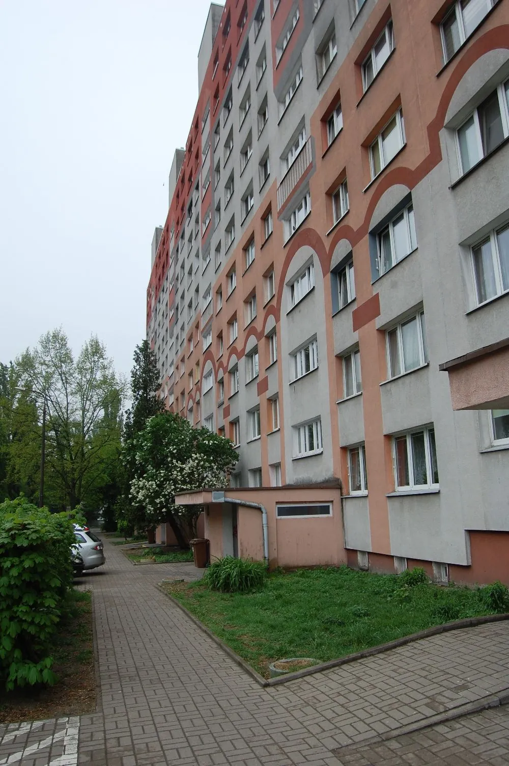 Wielopiętrowy blok mieszkalny o elewacji w kolorach szarym i pomarańczowym z licznymi oknami. Przed budynkiem znajduje się chodnik z kostki brukowej, trawnik oraz niskie, pomarańczowe przybudówki. Po lewej stronie widoczne są zielone krzewy, drzewa oraz zaparkowany srebrny samochód.