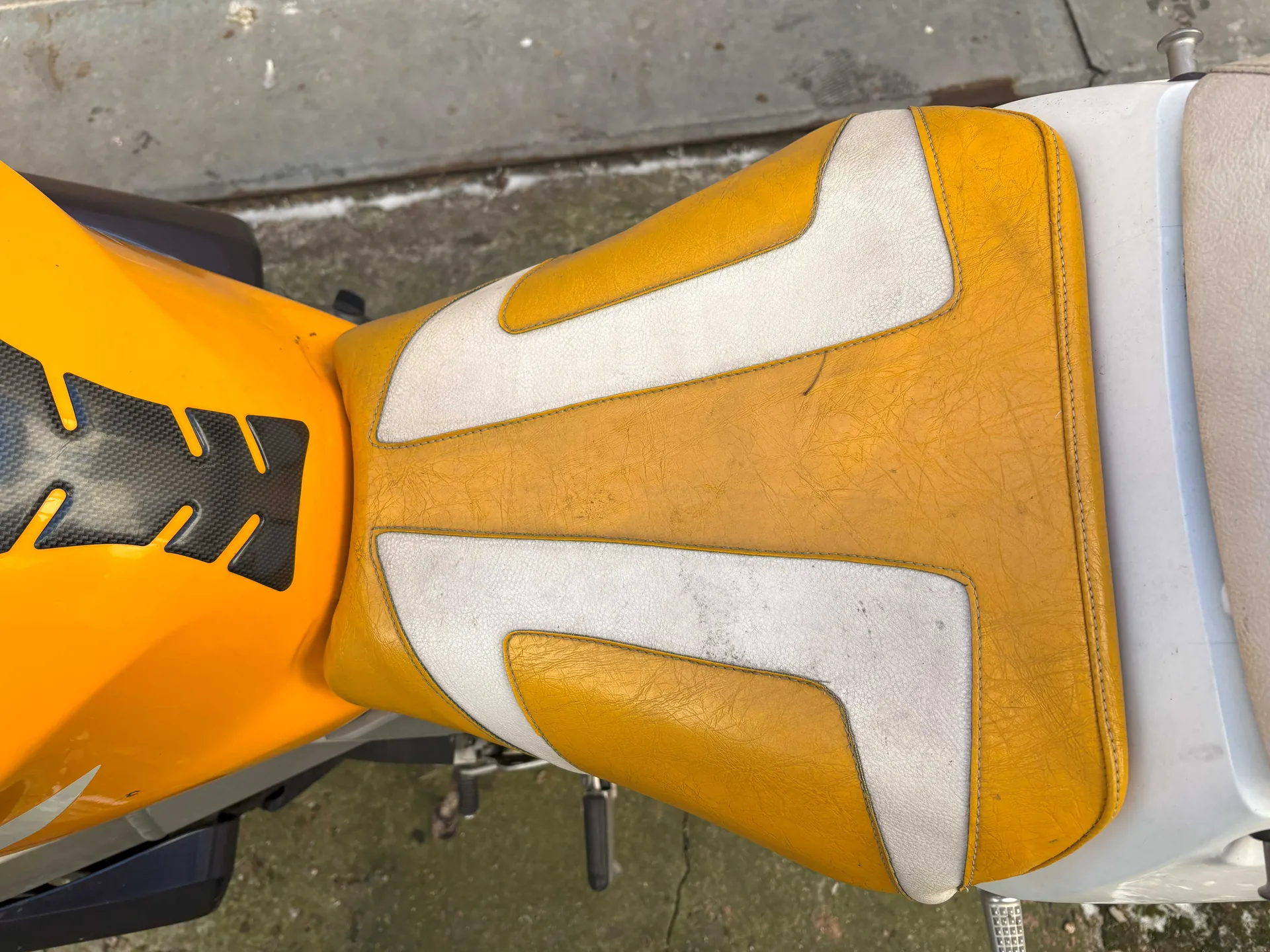 Zbliżenie na żółte siedzenie motocykla z srebrnymi wstawkami. Widoczna również żółta część zbiornika paliwa z czarnym wzorem przypominającym włókno węglowe i żółtymi akcentami. Tło stanowi betonowa powierzchnia i chodnik. Siedzenie posiada widoczne szwy i ślady zużycia.