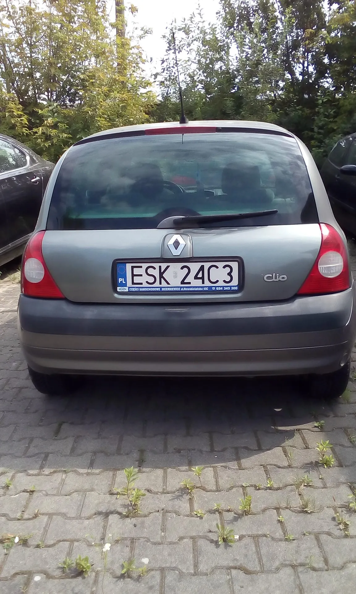 Tylny widok srebrnego samochodu marki Renault Clio zaparkowanego na kostce brukowej. Samochód ma polską tablicę rejestracyjną o numerze ESK 24C3. Po prawej stronie klapy bagażnika znajduje się napis 'Clio'. W tle widoczne są zielone drzewa i krzewy.
