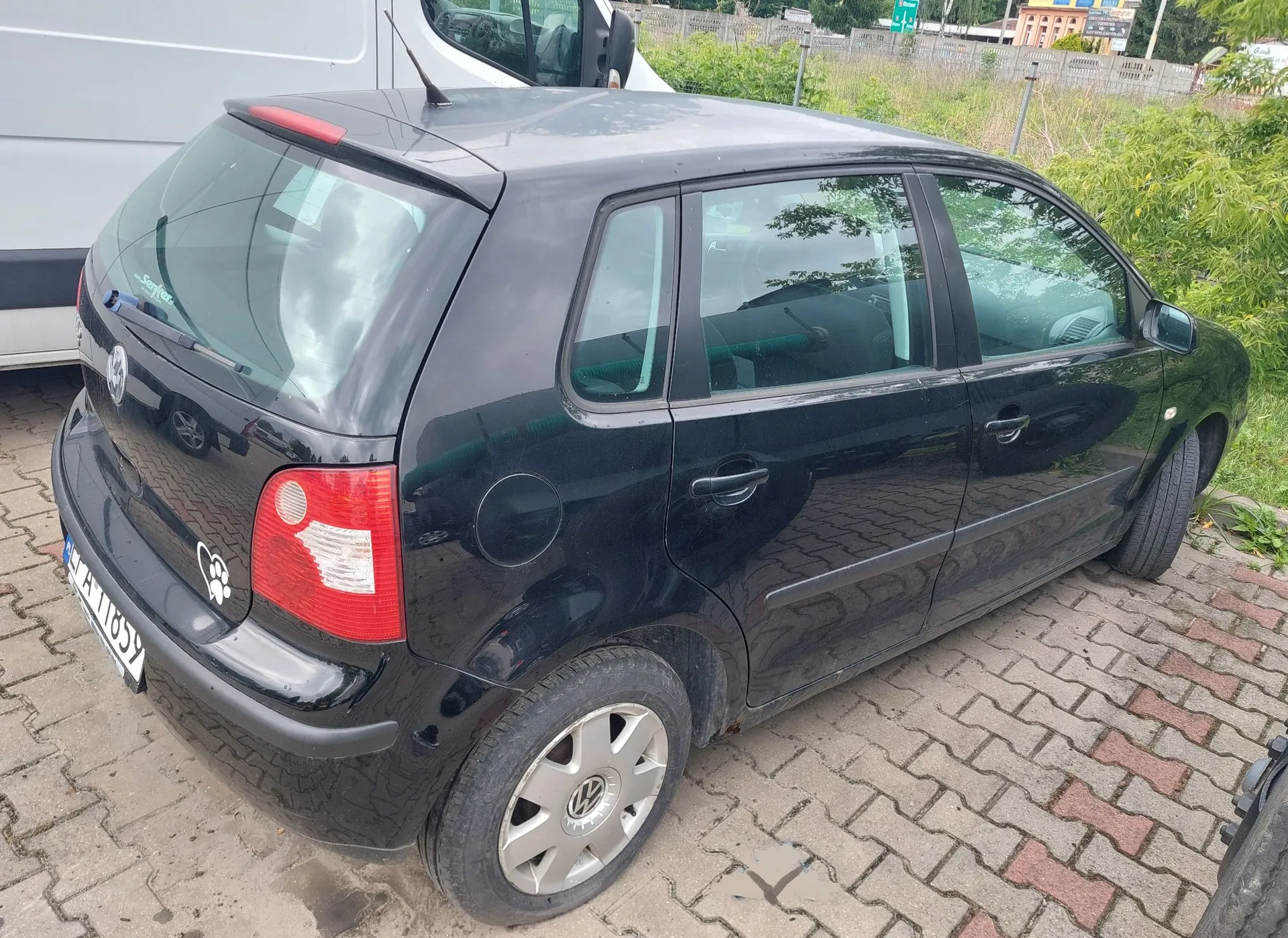 Czarny samochód marki Volkswagen Polo zaparkowany na kostce brukowej. Widok z tyłu i z boku auta. Na tylnej klapie znajduje się naklejka w kształcie białej łapy zwierzęcia oraz napis 'Safe Driver' na tylnej szybie. Samochód ma srebrne felgi aluminiowe z logo Volkswagena.