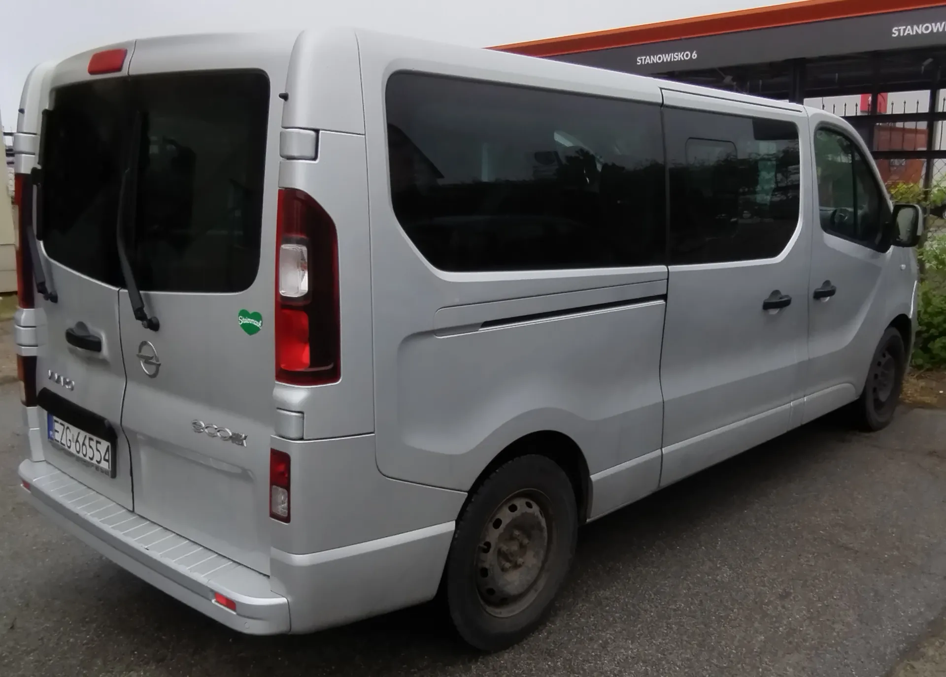 Srebrny samochód dostawczy marki Opel Vivaro zaparkowany na asfaltowej nawierzchni. Widok z tyłu i z boku pojazdu. Samochód ma przyciemniane szyby, tablicę rejestracyjną o numerze EZG 66554 oraz zieloną naklejkę w kształcie serca na tylnych drzwiach. W tle widoczny jest fragment budynku z pomarańczowym pasem i napisem 'STANOWISKO 6'.