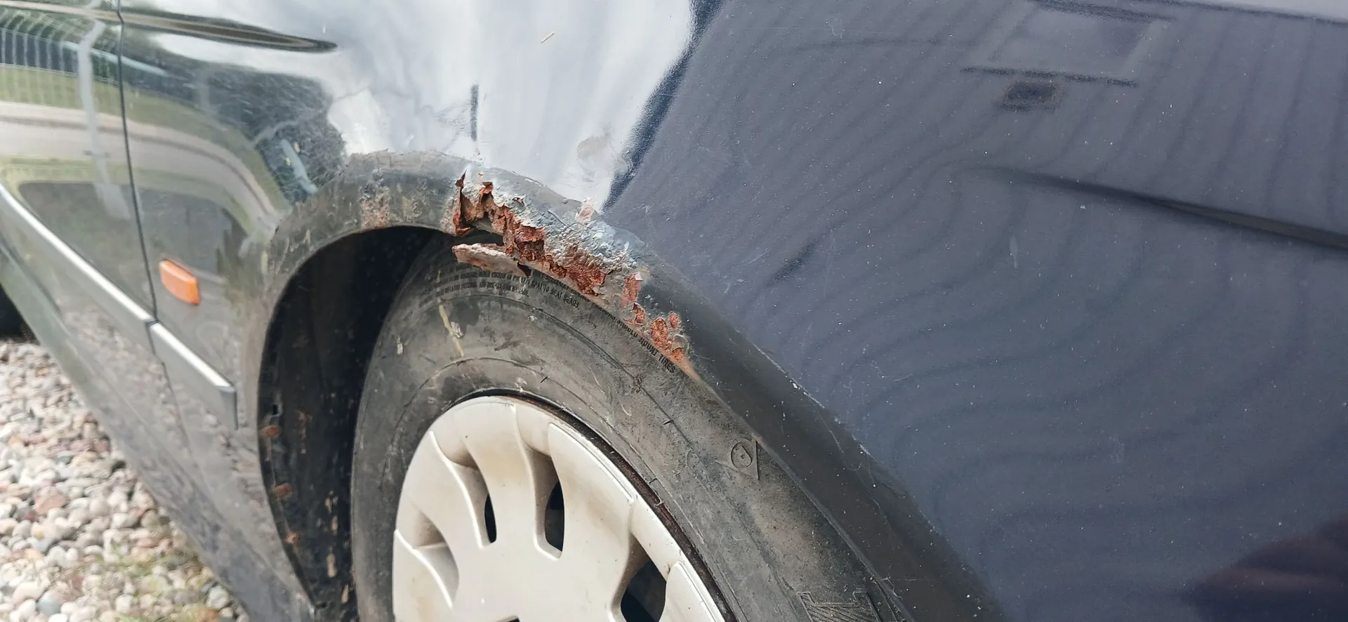 Zbliżenie na tylny błotnik ciemnoniebieskiego samochodu, w którym widoczne jest znaczne uszkodzenie karoserii w postaci głębokiej rdzy i ubytków w metalu nad nadkolem. Poniżej znajduje się opona z jasnym kołpakiem.