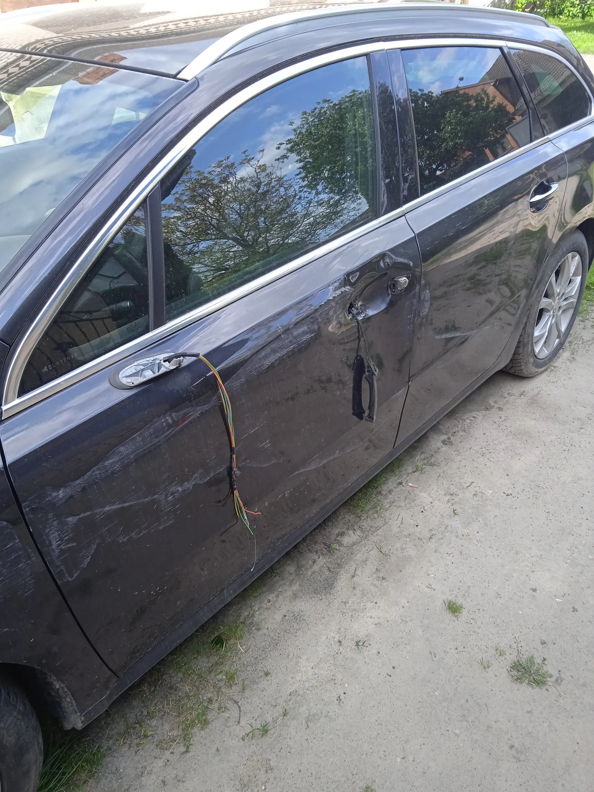 Czarny samochód typu kombi zaparkowany na betonowej powierzchni z rozproszoną trawą. Widoczne są zadrapania na przedniej drzwi oraz kolorowe przewody wiszące przy uchwycie drzwi. W szybach odbijają się drzewa i budynki. Samochód stoi na tle ziemi z rzadką trawą.
