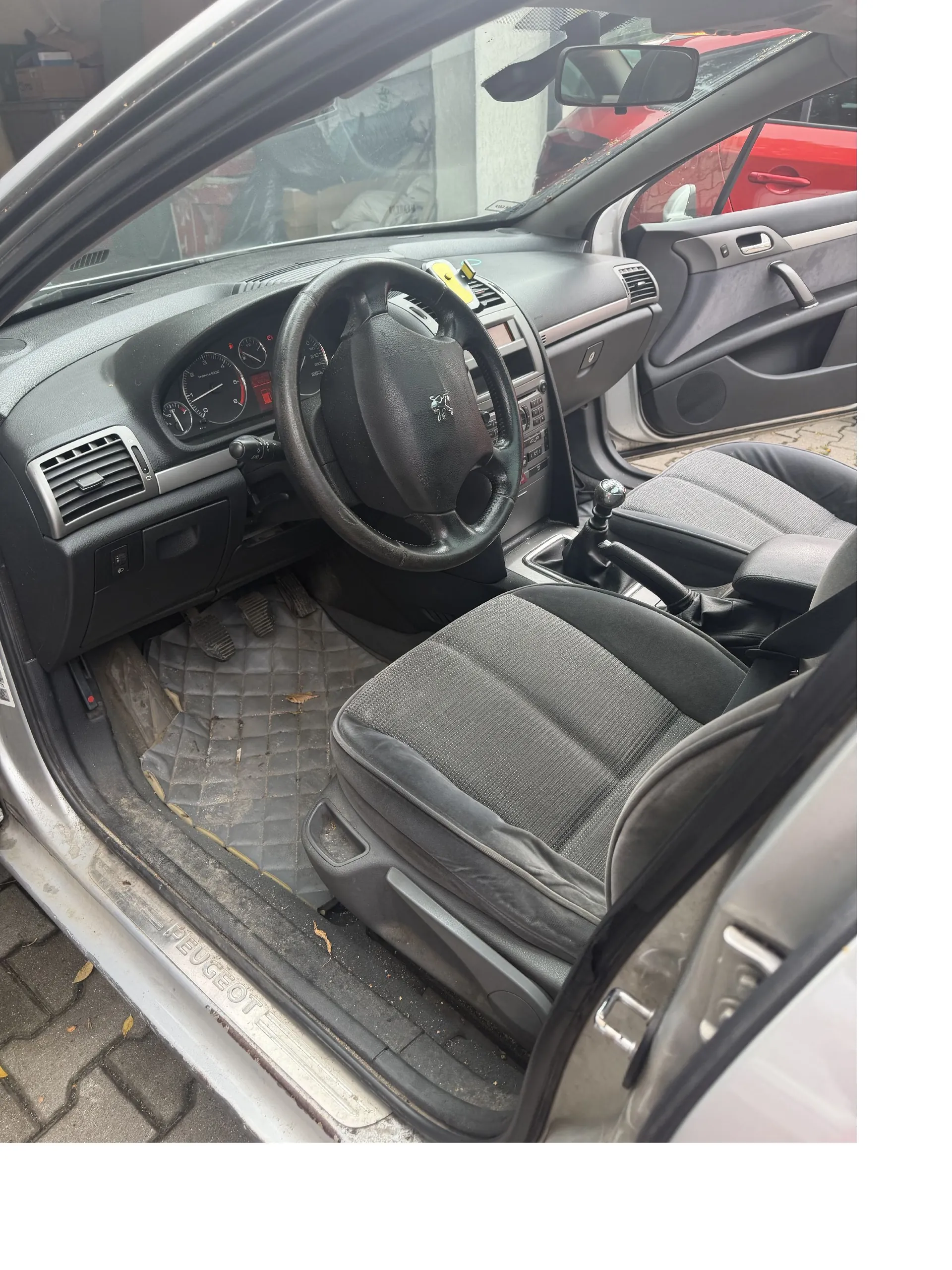Wnętrze srebrnego samochodu marki Peugeot widziane z otwartych drzwi kierowcy. Widoczna jest czarna kierownica z logo marki, deska rozdzielcza z zegarami, lewy fotel z ciemnoszarą tapicerką oraz dywanik z rombowym wzorem. Na progu samochodu znajduje się napis 'PEUGEOT'. Przez szybę w tle widoczny jest fragment czerwonego samochodu.