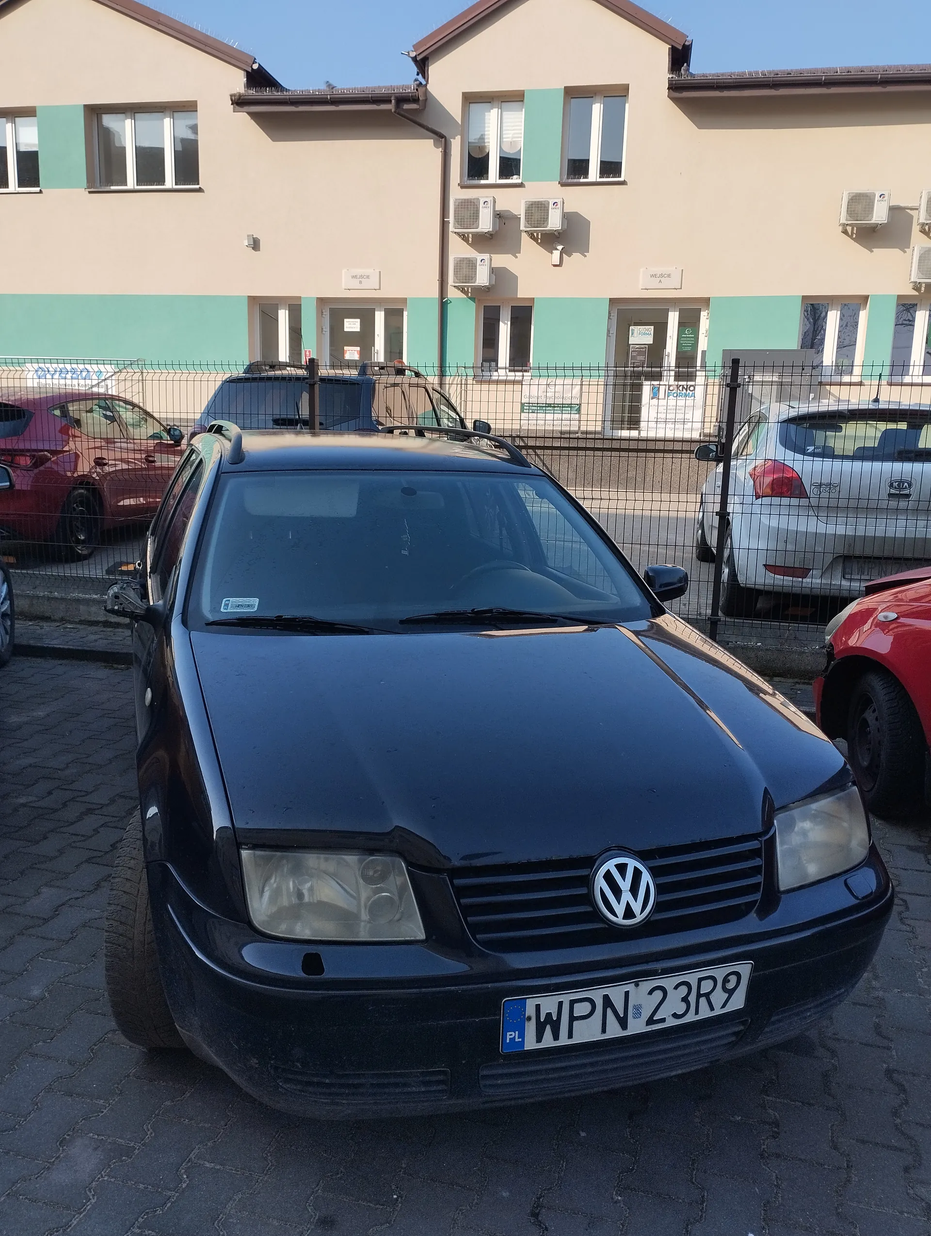 Czarny samochód marki Volkswagen z tablicą rejestracyjną WPN 23R9 (PL) zaparkowany na brukowanym placu. W tle budynki o beżowo-turkusowej elewacji z oknami i klimatyzatorami, metalowe ogrodzenie oraz inne zaparkowane pojazdy: czerwony, srebrny i czarny. Niebo jest bezchmurne.
