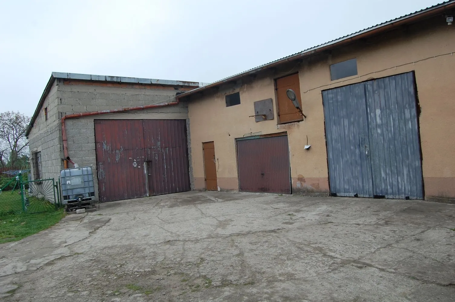 Widok na stare, gospodarcze budynki z betonowego i tynkowanego muru. Po lewej znajduje się szary budynek z dużymi, brązowymi, metalowymi drzwiami garażowymi oraz plastikowy zbiornik IBC. Po prawej znajduje się dłuższy budynek o beżowej elewacji z dwiema bramami: jedną mniejszą, brązową oraz jedną większą, szarą, wykonaną z pionowych blach. Przed budynkami znajduje się betonowy plac z pęknięciami.