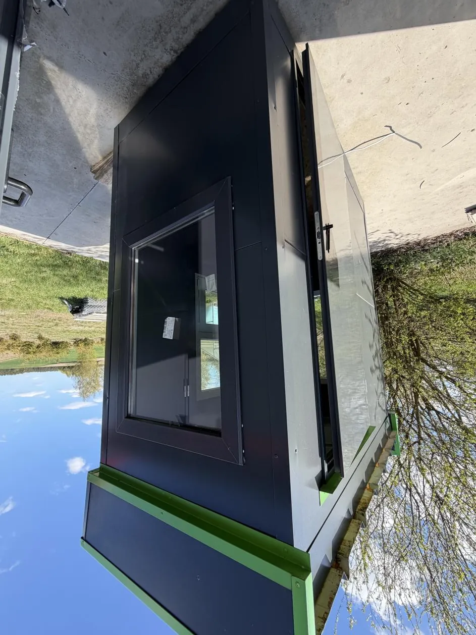 Czarny, prostokątny obiekt z zielonymi akcentami na krawędziach, przypominający małą budkę lub kiosk. Obiekt posiada duże okno z czarną ramą i częściowo otwarte drzwi. W tle widoczne są betonowa powierzchnia, trawa, drzewa oraz niebo z białymi chmurami. Na szybie okna znajduje się mały, biały dokument lub naklejka.
