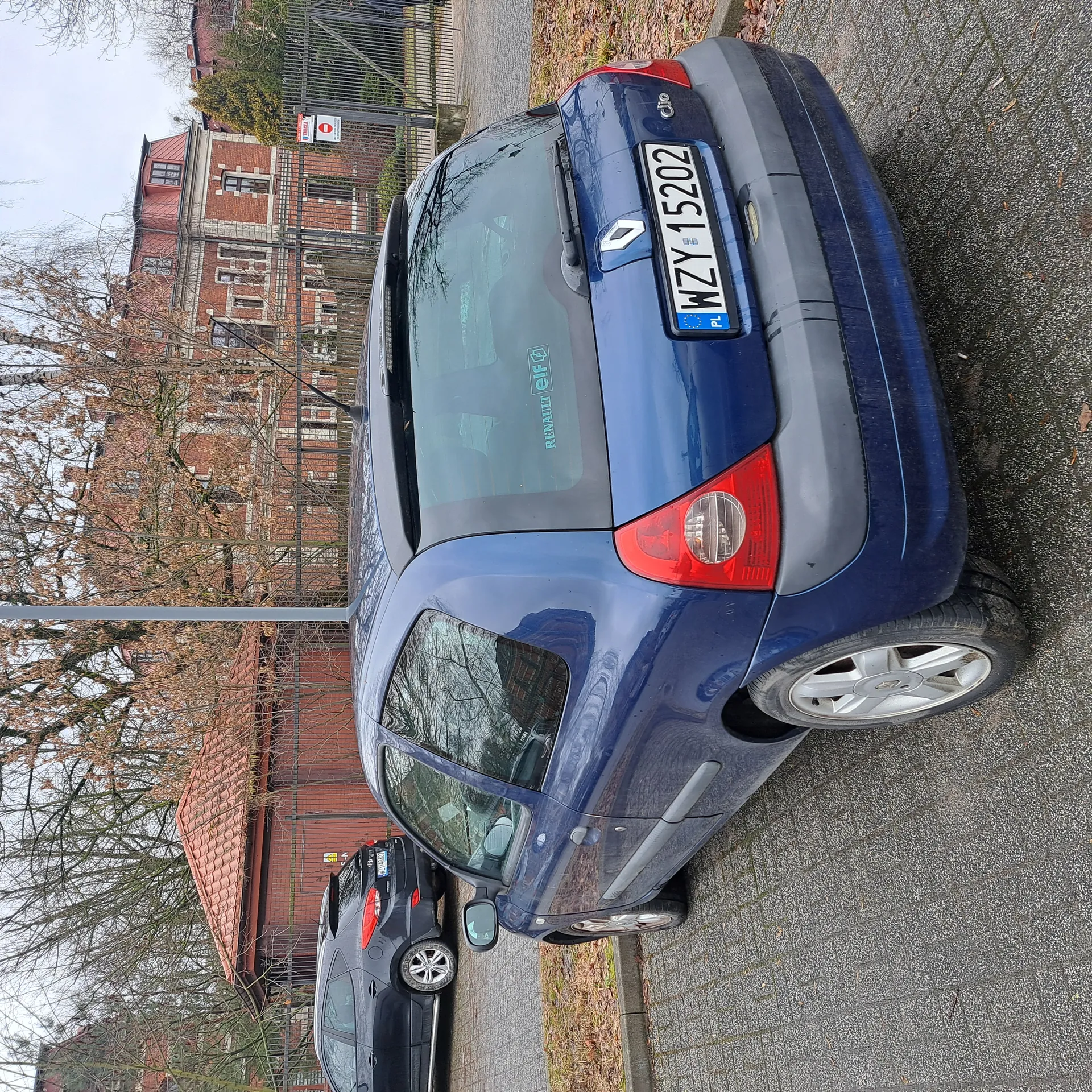 Niebieski samochód Renault Clio zaparkowany tyłem do obserwatora na brukowanym placu. Widoczna tablica rejestracyjna PL WZY 15202 oraz naklejka 'RENAULT elf' na tylnej szybie. W tle czarny samochód, czerwona cegłana budowla z dachem krytym dachówką, metalowe ogrodzenie oraz większy budynek z czerwonym dachem. Drzewa bez liści, na ziemi suche liście. Znak zakazu wjazdu przy ogrodzeniu.
