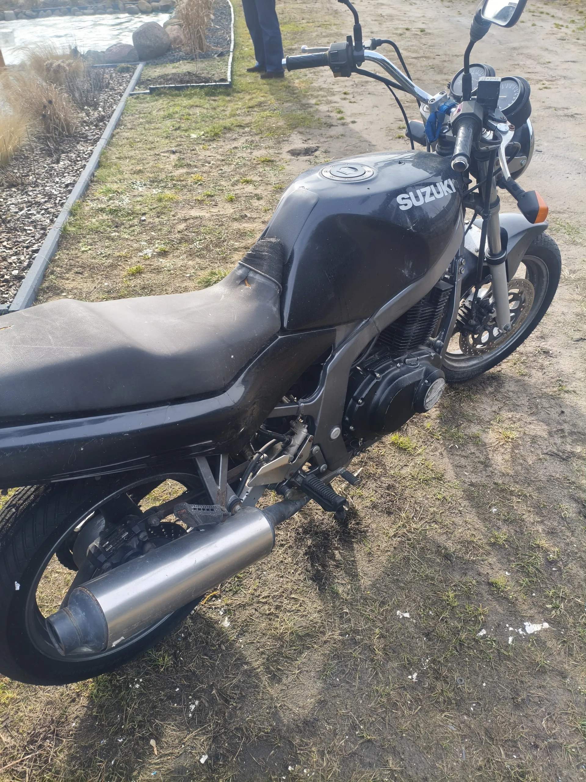 Czarny motocykl marki SUZUKI z widocznym napisem na zbiorniku paliwa, stojący na trawiastym i częściowo ziemistym terenie. Widoczne są czarne siedzenie z uszkodzeniem, wydech, kierownica z lusterkami oraz wskaźnikami. W tle fragment ogrodu z kamieniami, suchą trawą, niewielkim ciekem wodnym oraz nogami stojącej osoby w ciemnych spodniach i butach.