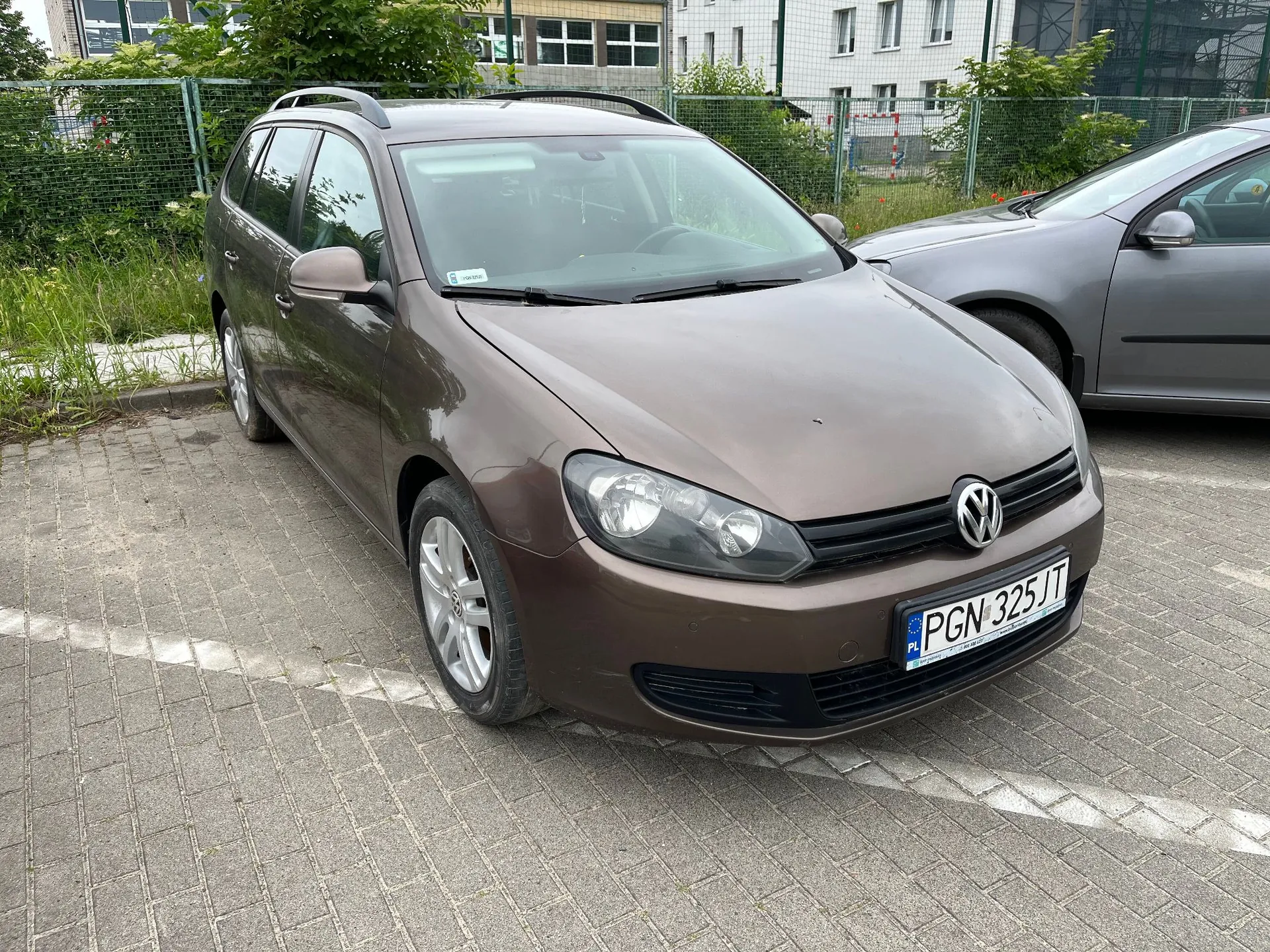 Brązowy samochód osobowy marki Volkswagen Golf w wersji kombi, zaparkowany na kostce brukowej. Pojazd ma polską tablicę rejestracyjną o numerze PGN 325JT. W tle widoczne są zielone krzewy, ogrodzenie z siatki oraz biały budynek z oknami. Obok stoi fragment innego, szarego samochodu.