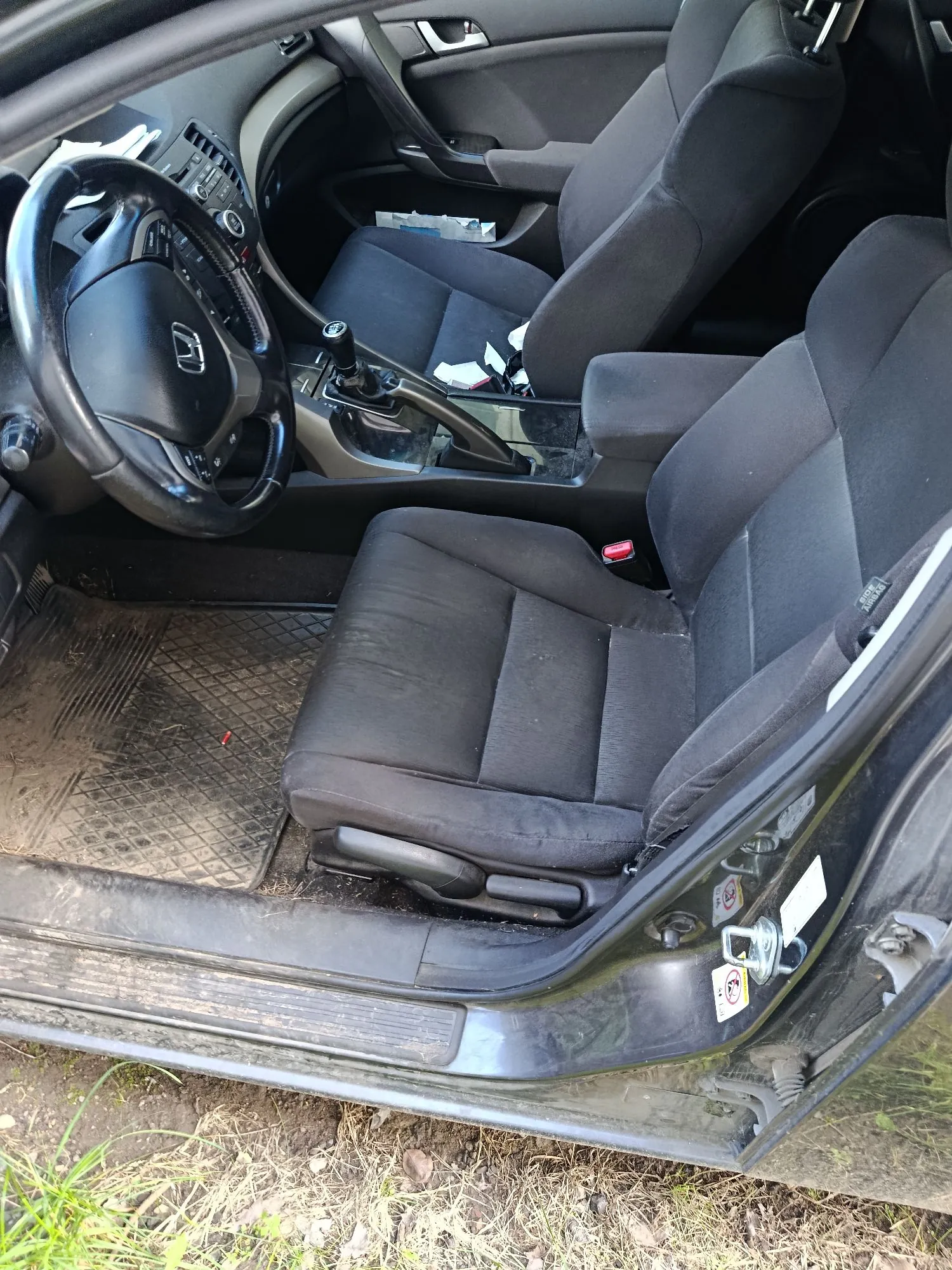 Wnętrze samochodu widziane z otwartych drzwi pasażera. Widoczna jest czarna tapicerka materiałowa na przednich fotelach, czarna kierownica z logo marki Honda, konsola środkowa z lewarkiem zmiany biegów oraz brudna mata podłogowa po stronie kierowcy. Wewnątrz pojazdu, na siedzeniach i podłodze, znajdują się rozrzucone białe papiery.