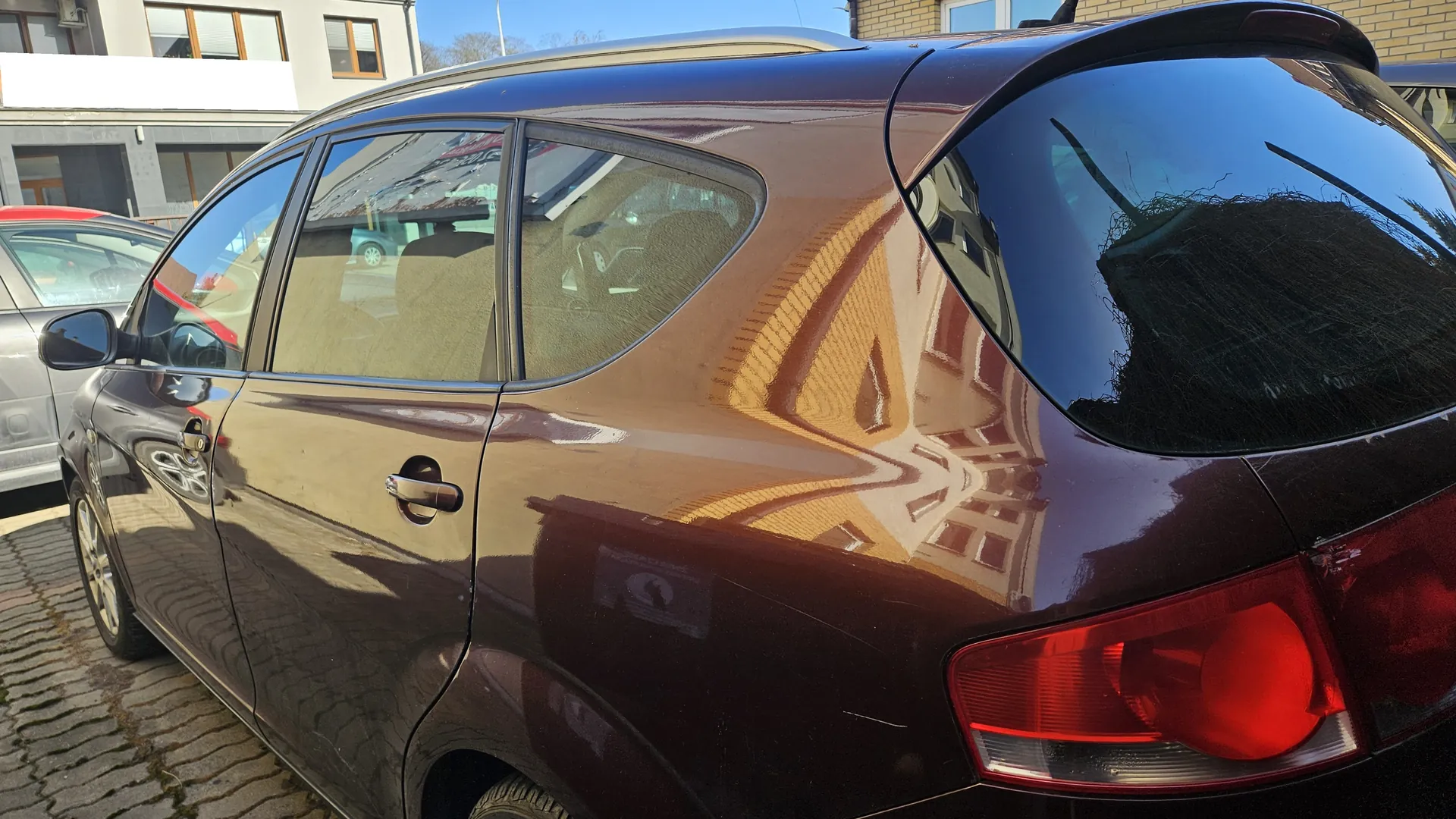 Ciemnobrązowy samochód osobowy typu hatchback zaparkowany na brukowanej ulicy. Widok z tyłu i boku. W tle inne pojazdy, budynki z oknami oraz jasne niebo. Na szybach odbijają się otoczenie i budynki.