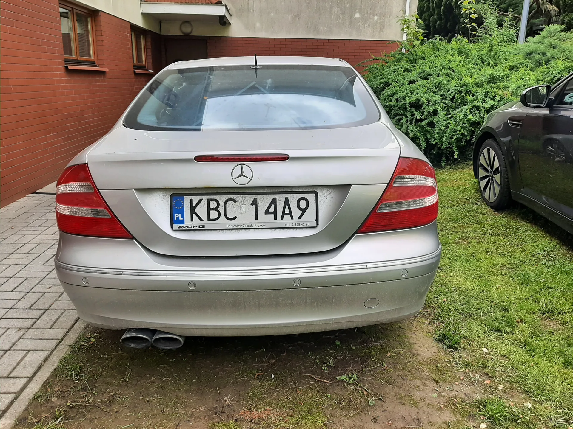 Z tyłu srebrny samochód marki Mercedes-Benz z tablicą rejestracyjną KBC 14A9. Na lewej stronie tablicy widoczny jest niebieski pas z literą PL i flagą Unii Europejskiej. Poniżej tablicy znajduje się napis 'AMG' oraz 'Sobiesław Zasada Kraków' oraz numer telefonu 'tel. 12 298 42 20'. Samochód stoi na trawie obok czerwonej ceglanej ściany budynku z oknami. Po prawej stronie częściowo widoczny jest czarny samochód. Tło stanowią zielone krzewy i fragment szarej elewacji budynku.