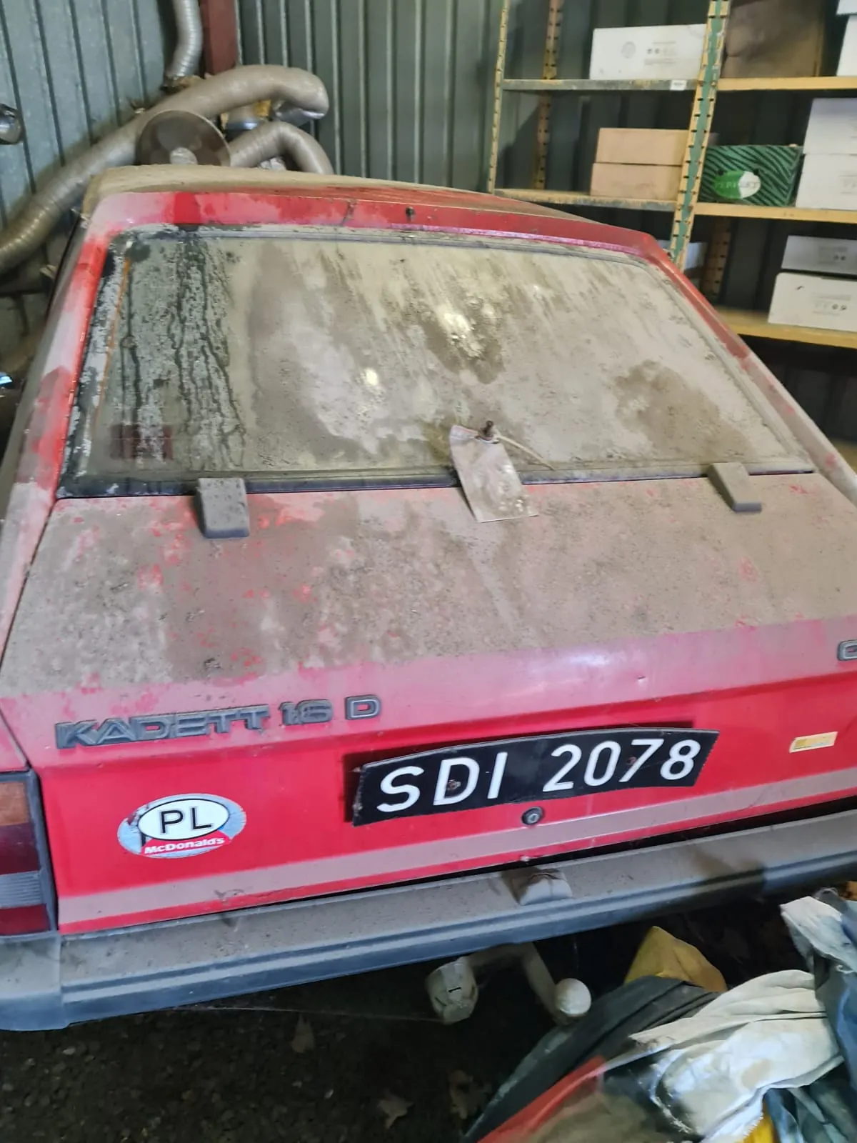 Czerwony samochód Opel Kadett 16 D z tyłu, pokryty kurzem i brudem. Widoczny tabliczka rejestracyjna SDI 2078, naklejka McDonald's z napisem PL oraz napis KADETT 16 D na bagażniku. Tło stanowi garaż z metalowymi półkami, na których znajdują się pudełka i opakowania, oraz rury wentylacyjne przy ścianie.
