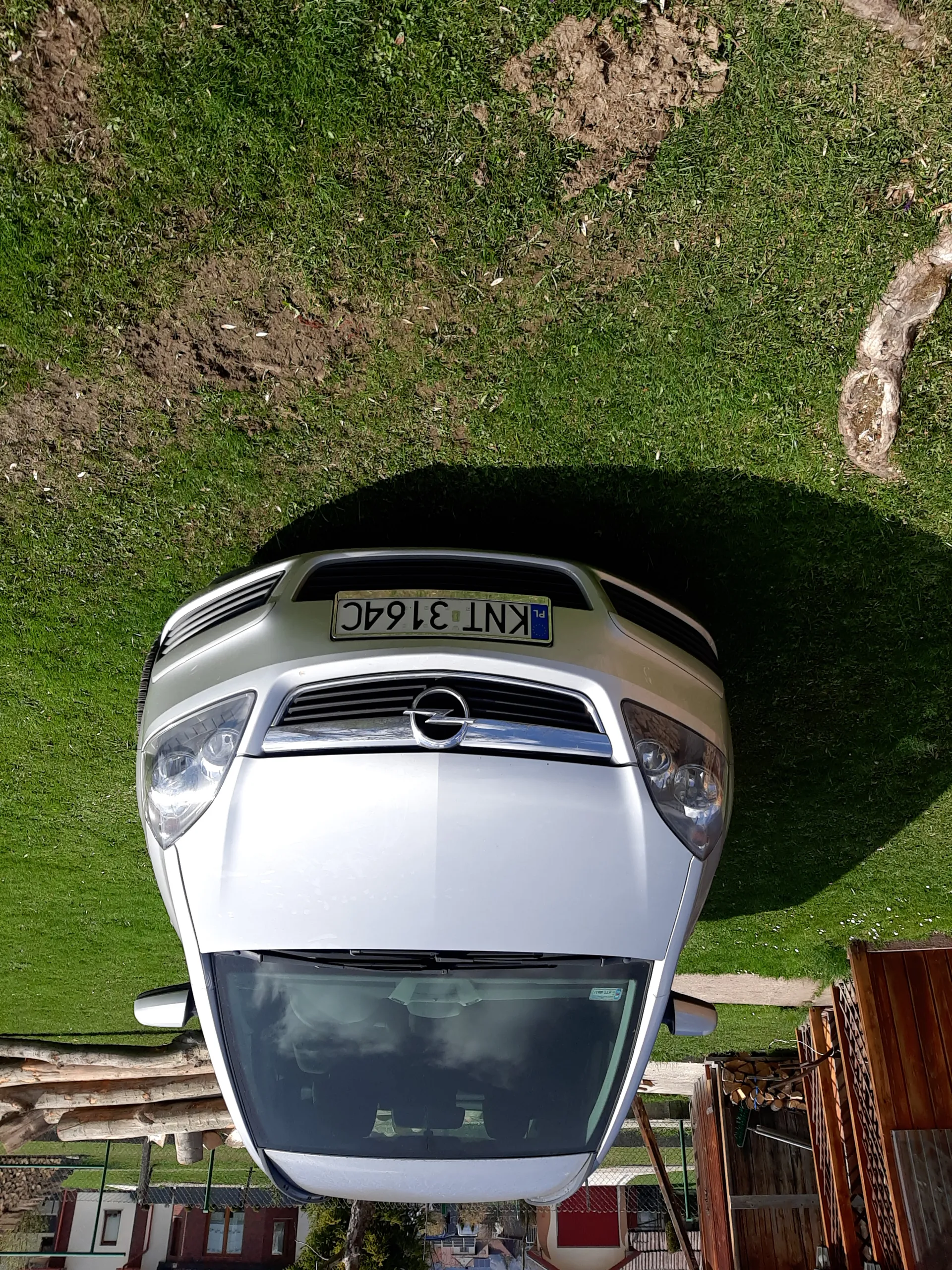 Srebrny samochód marki Opel z pionowo umieszczoną tablicą rejestracyjną KNT 3164C zaparkowany na zielonej trawie. W tle widoczne są drewniane konstrukcje, stosy drewna, domy oraz drzewa.