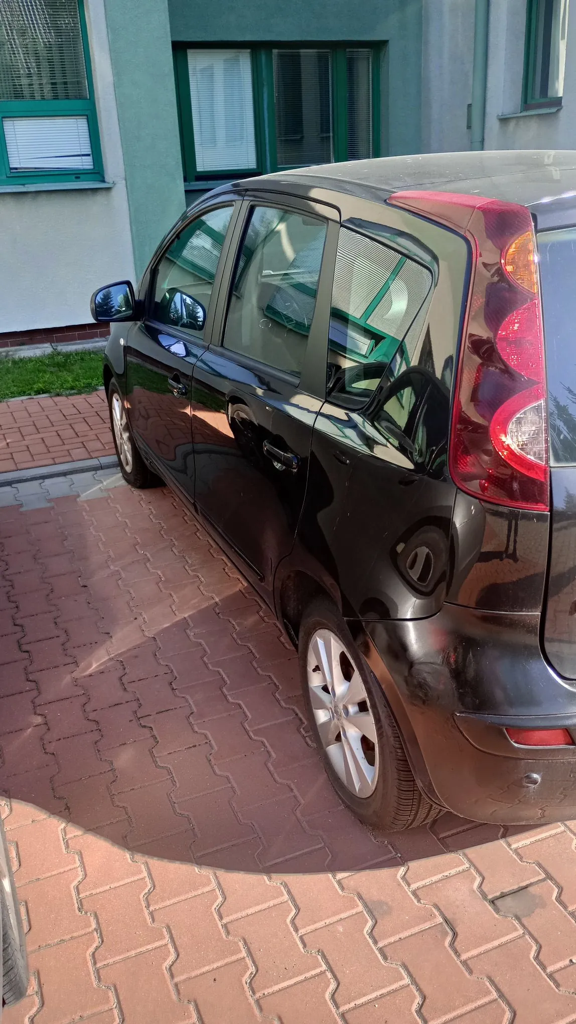 Czarny samochód osobowy zaparkowany na czerwonym kostkowym podjeździe przed zielonym budynkiem. Widoczny jest tył i bok auta, w tym tylna lampa, koła oraz szyby, w których odbija się otoczenie.