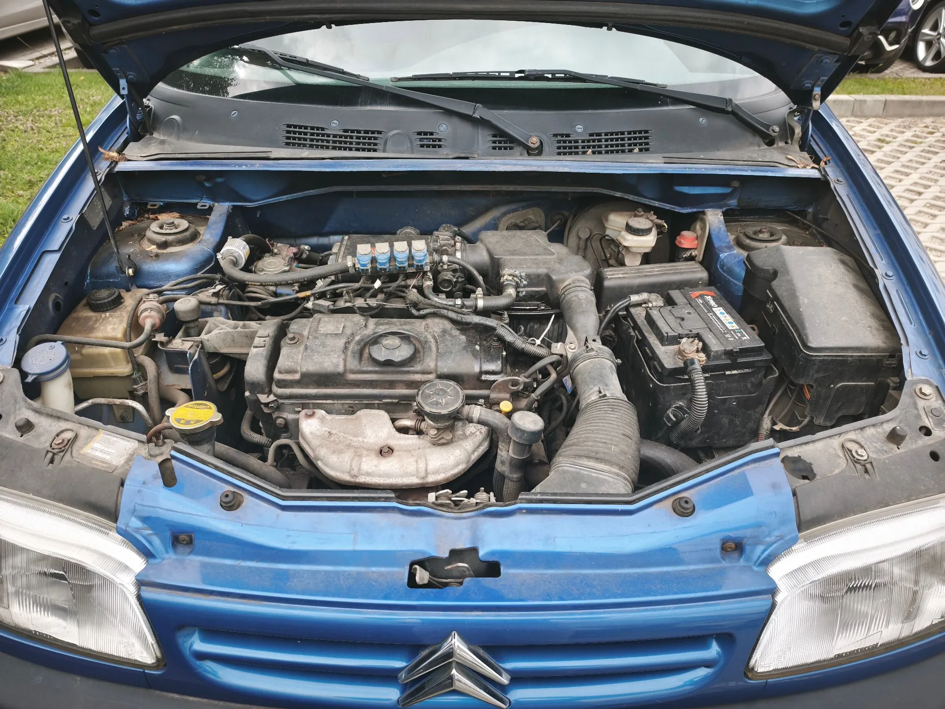Widok z góry na otwartą maskę niebieskiego samochodu marki Citroën, ukazującą komorę silnika. Widoczny jest silnik spalinowy, akumulator po prawej stronie, zbiorniki z płynami eksploatacyjnymi oraz elementy układu chłodzenia i elektryki. Na przodzie maski widoczne jest logo Citroëna.