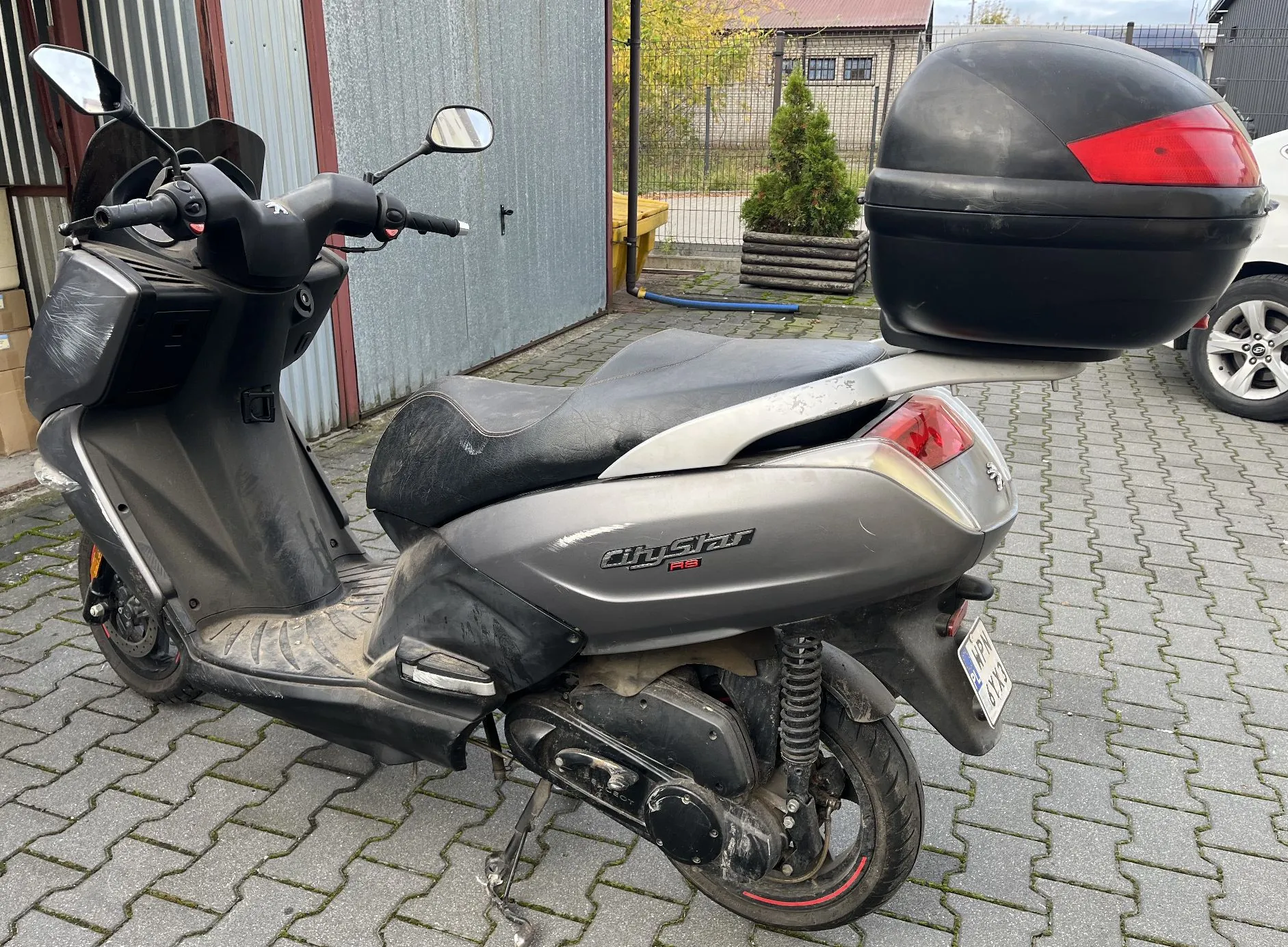 Srebrny skuter marki CityStar RS zaparkowany na kostce brukowej. Pojazd posiada czarny kufer zamykany z tyłu, czarną kanapę oraz lusterka boczne. Na tylnym błotniku widoczna jest tablica rejestracyjna. W tle znajduje się szary budynek z blachy falistej oraz mały zielony krzew w donicy.