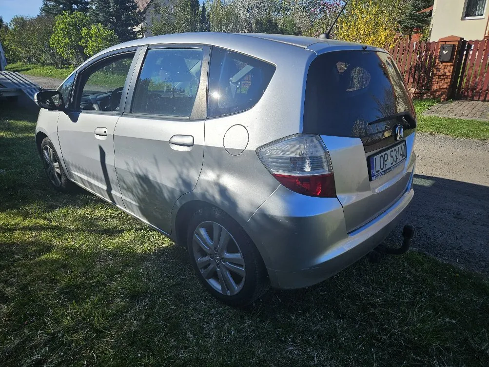 Srebrny samochód osobowy marki Honda widziany z tyłu i boku, zaparkowany na trawie. Widoczna tablica rejestracyjna LOP 53140, tylne światła, felgi oraz otoczenie z domem, czerwonym ogrodzeniem i drzewami. W tle zielone krzewy i niebo.