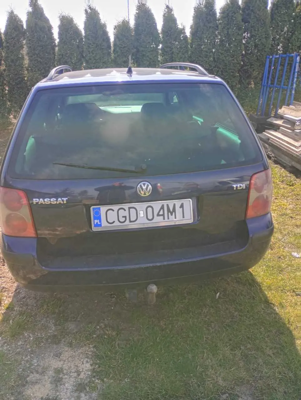 Czarny samochód osobowy Volkswagen Passat TDI z widocznym tyłem. Na bagażniku umieszczono napisy 'PASSAT' po lewej stronie i 'TDI' po prawej stronie. W centrum bagażnika znajduje się logo Volkswagen. Tablica rejestracyjna oznaczona jako 'CGD 04M1' z niebieskim pasem po lewej stronie zawierającym literę 'PL' i symbol Unii Europejskiej. Samochód stoi na trawie z fragmentem żwiru, na tle rzędu wysokich, zielonych drzew iglastych. Po prawej stronie widoczna jest niebieska metalowa konstrukcja oraz stosy drewnianych desek.