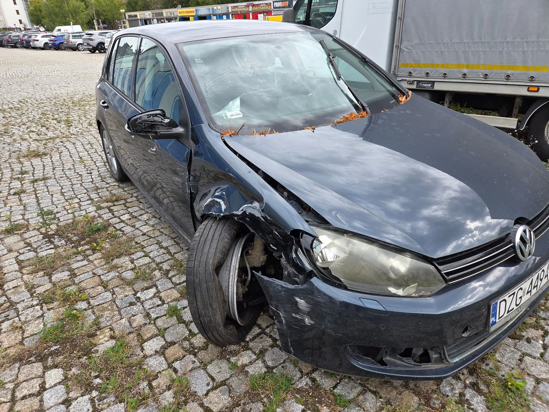 Ciemnoniebieski samochód marki Volkswagen Golf z poważnymi uszkodzeniami przodu po prawej stronie. Przednie prawe koło jest wygięte i nienaturalnie odchylone, błotnik jest wgnieciony, a maska lekko podniesiona. Pojazd stoi na brukowanym placu. W tle widoczne są inne zaparkowane samochody oraz biała ciężarówka. Na zderzaku widoczna jest tablica rejestracyjna o numerze DZG 449A.